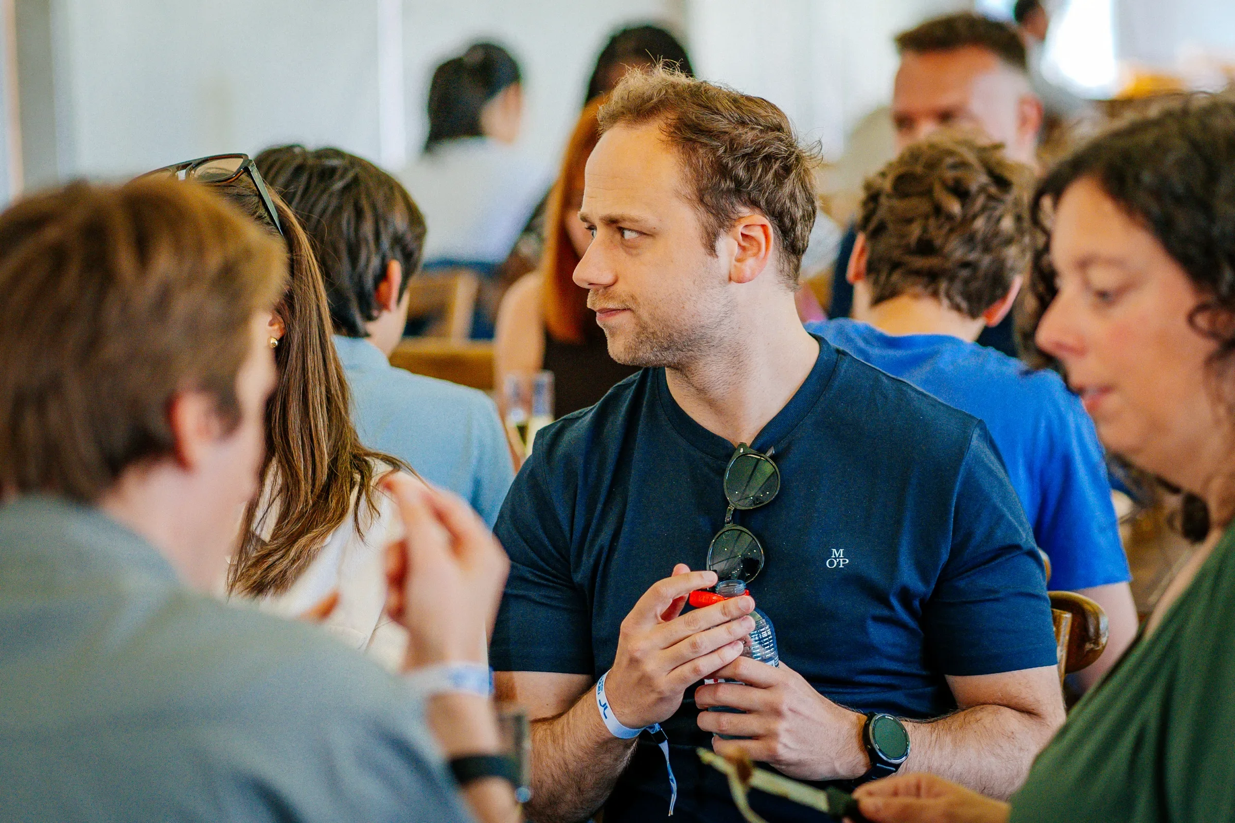 Man in blauwe T-shirt met zonnebril in de nek, omringd door andere mensen, waarbij hij een fles water vasthoudt, in een binnenruimte tijdens een sociale bijeenkomst of evenement.