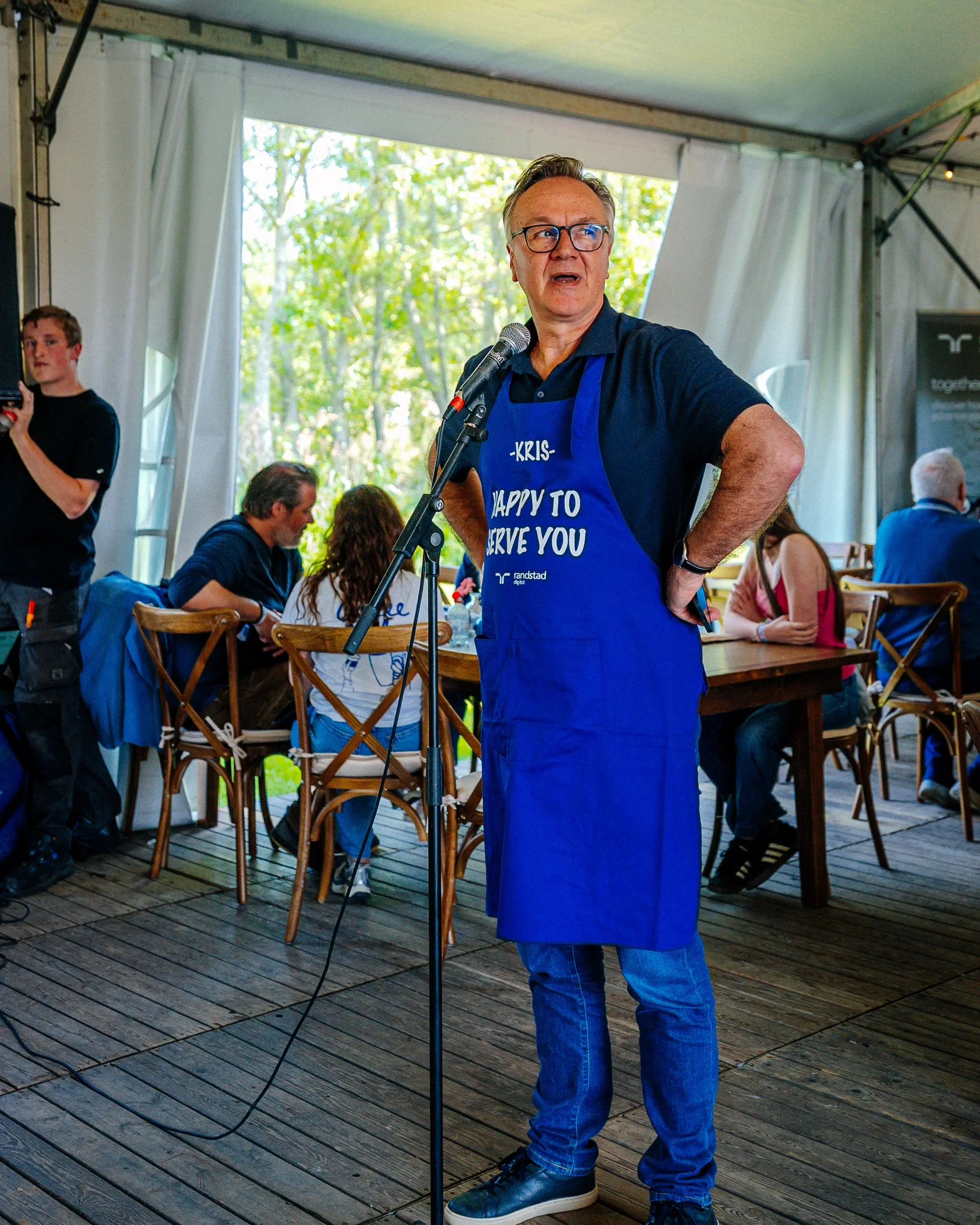 Man met bril, blauwe schort met tekst 'KRIS, HAPPY TO SERVE YOU', staat achter microfoon, binnen, met open tent en grote ramen met uitzicht op groen buiten, omringd door andere mensen.