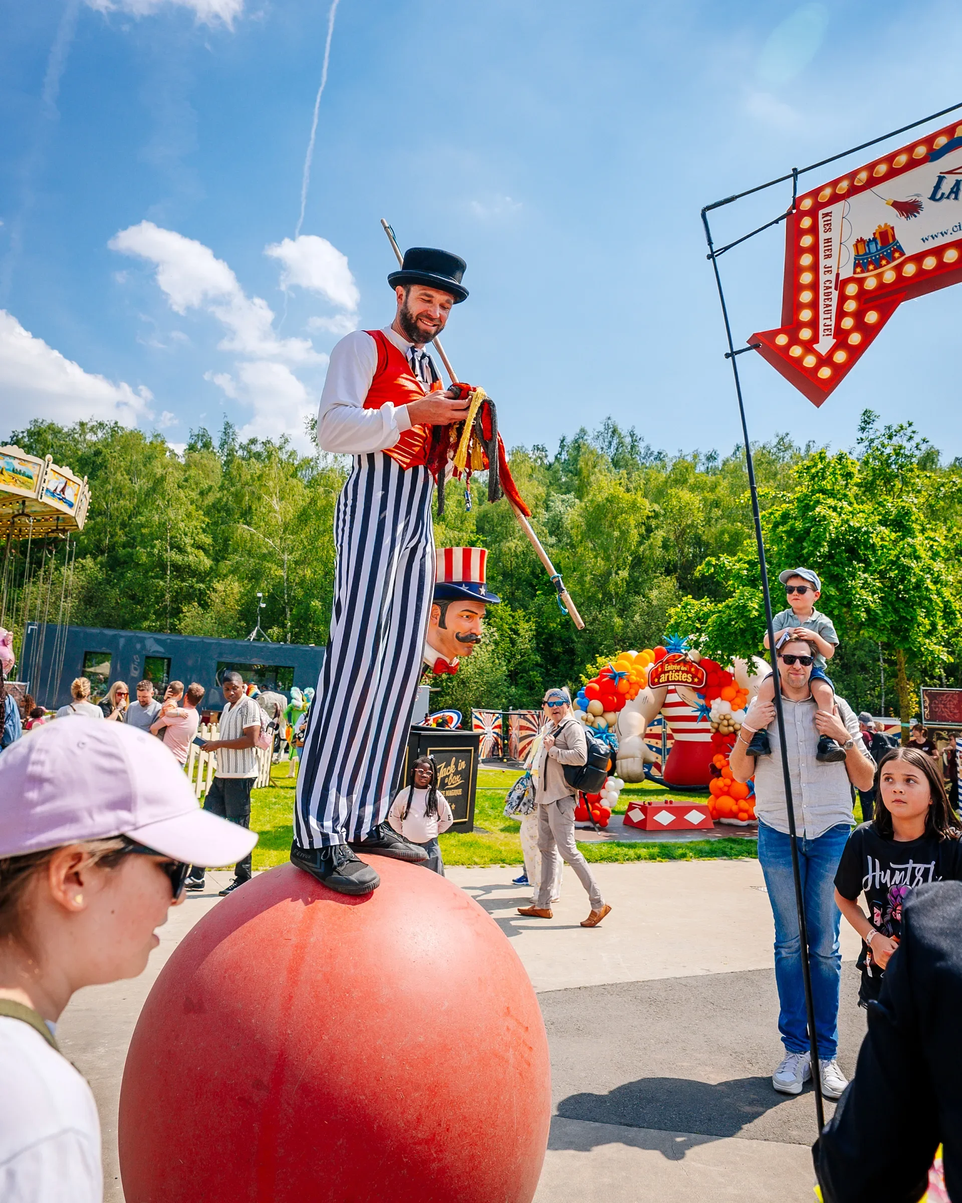 Man met circusoutfit en een zwarte hoed staat op een grote rode bal tijdens een kermis of markt, omgeven door mensen, met een attractie en kleurrijke ballonnen op de achtergrond.