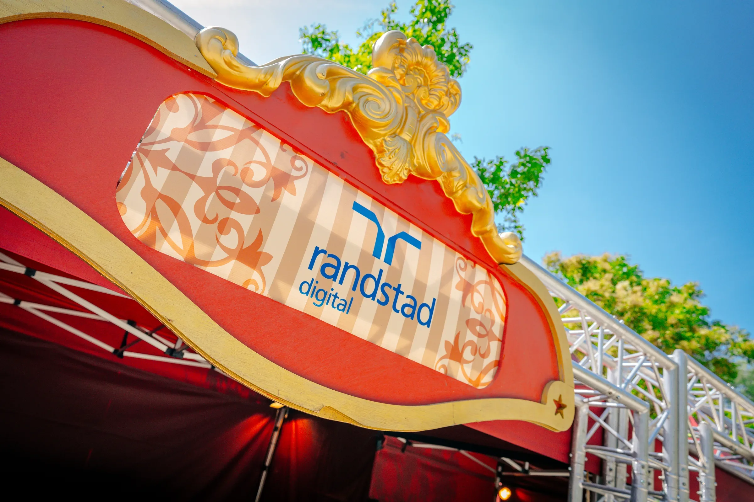 etailage van een attractie in een pretpark met een rode en gele decoratie en een blauw kruislogo, onder een blauwe lucht en groene bomen.