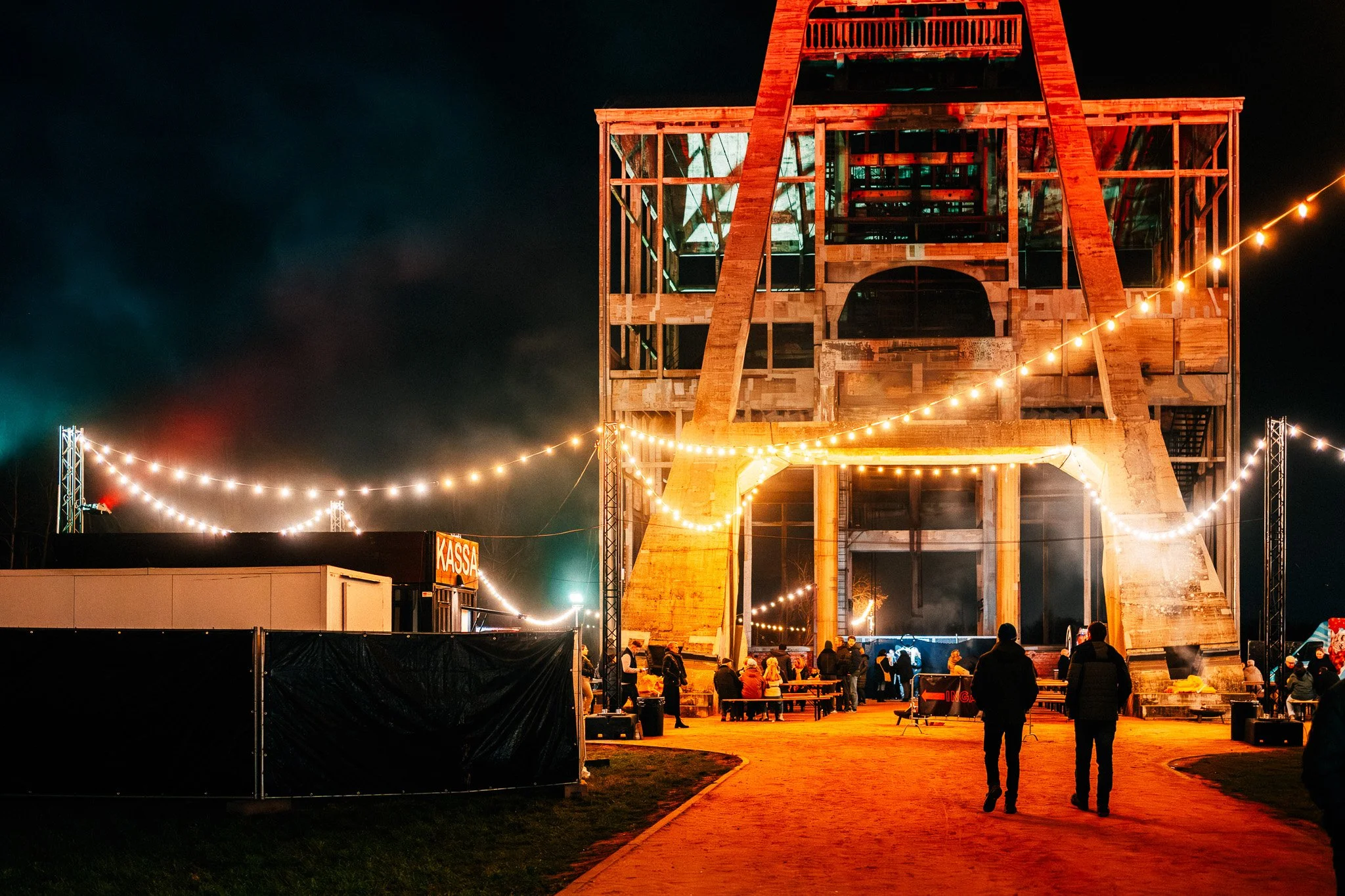 Een gebouw onder constructie verlicht met gekleurd licht, met mensen die erbuiten staan en genieten van het uitzicht, een gezellige sfeer met kerstlichten.