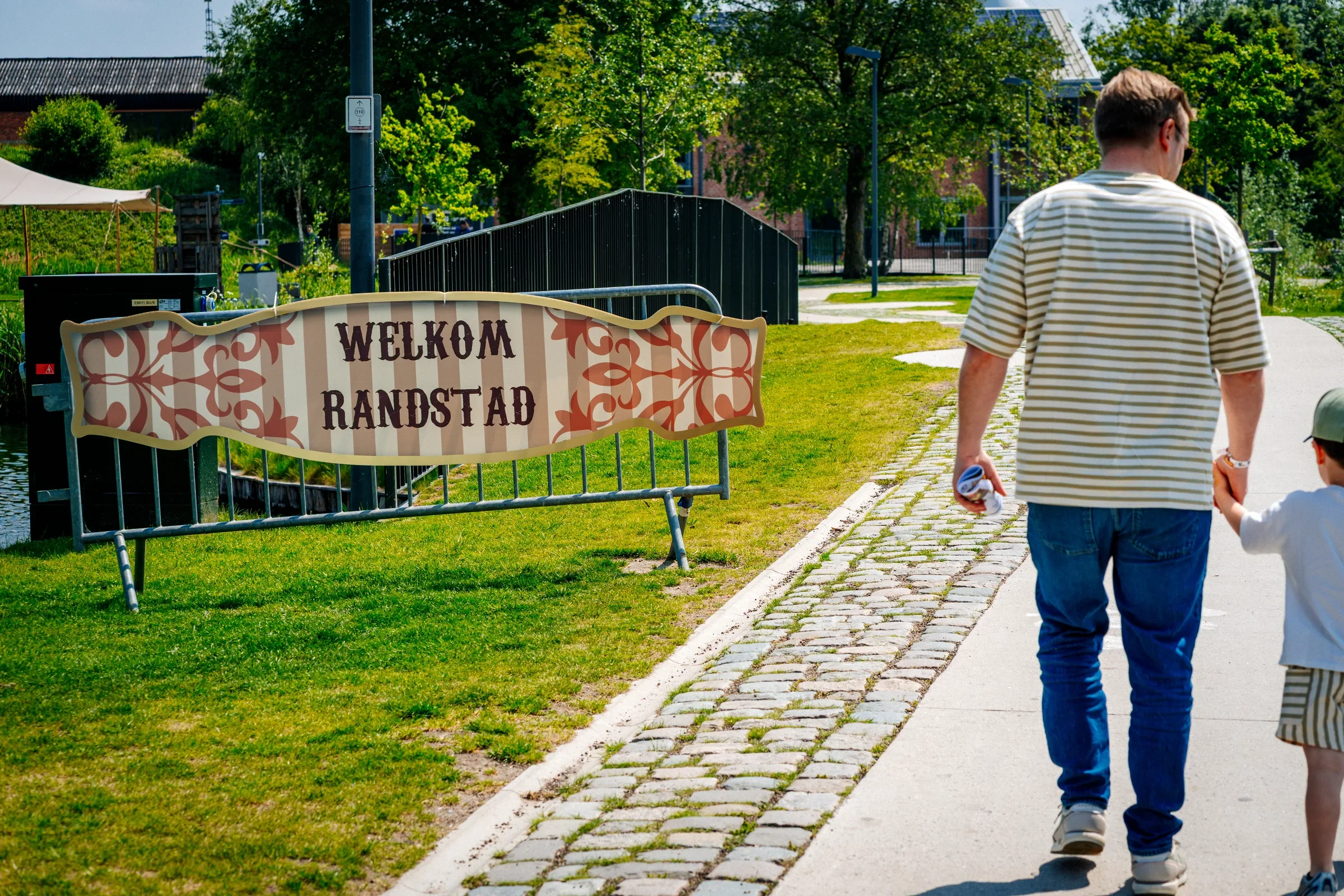 Man en kind lopen hand in hand langs een groen park met een hek en een horecagelegenheid. Er staat een welkomstbord met de tekst 'Welkom Randstad'.