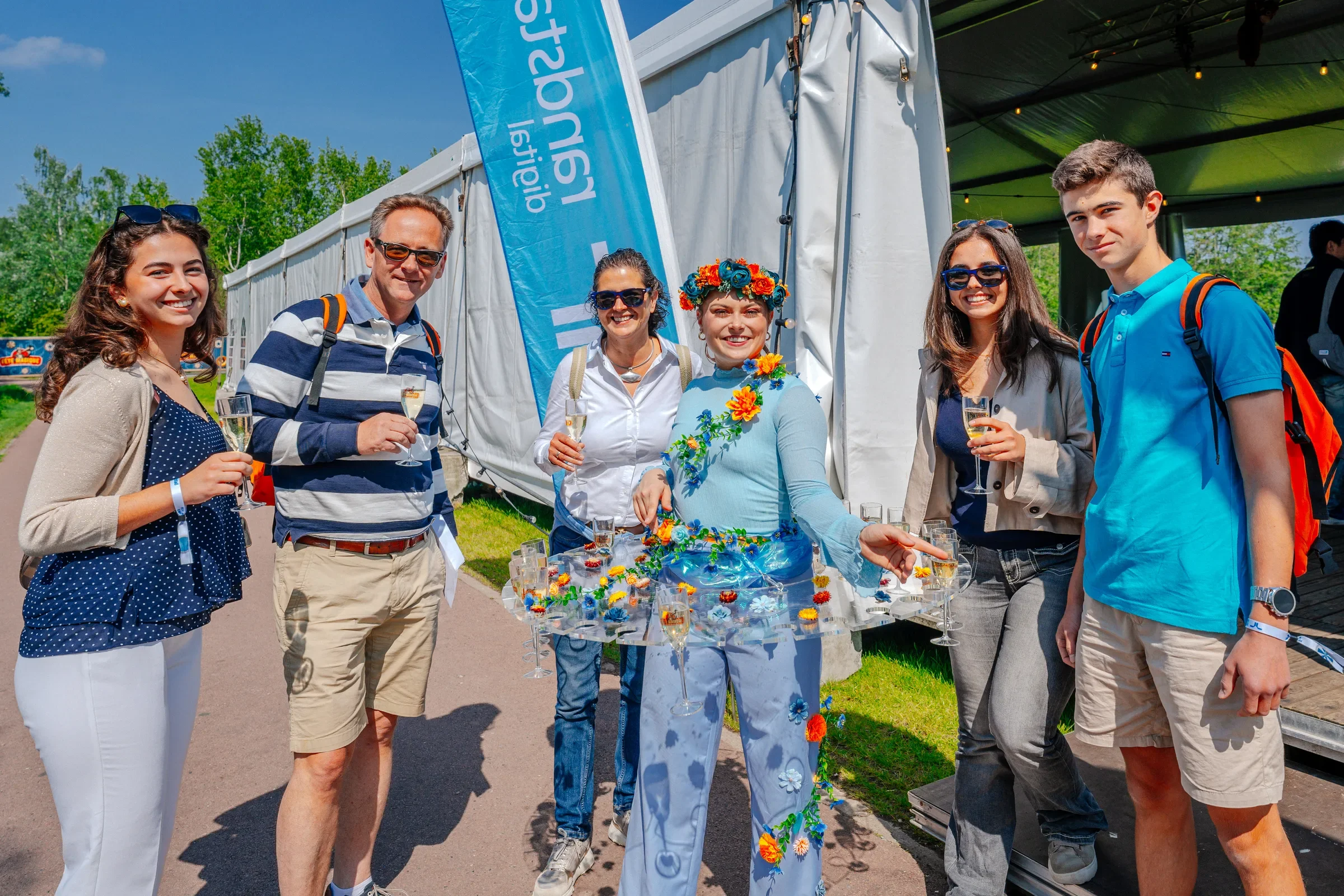 Vijf mensen staan in een groep buiten op een zonnige dag, enkele met glazen met champagne, voor een stand met bloemen en een blauwe vlag.