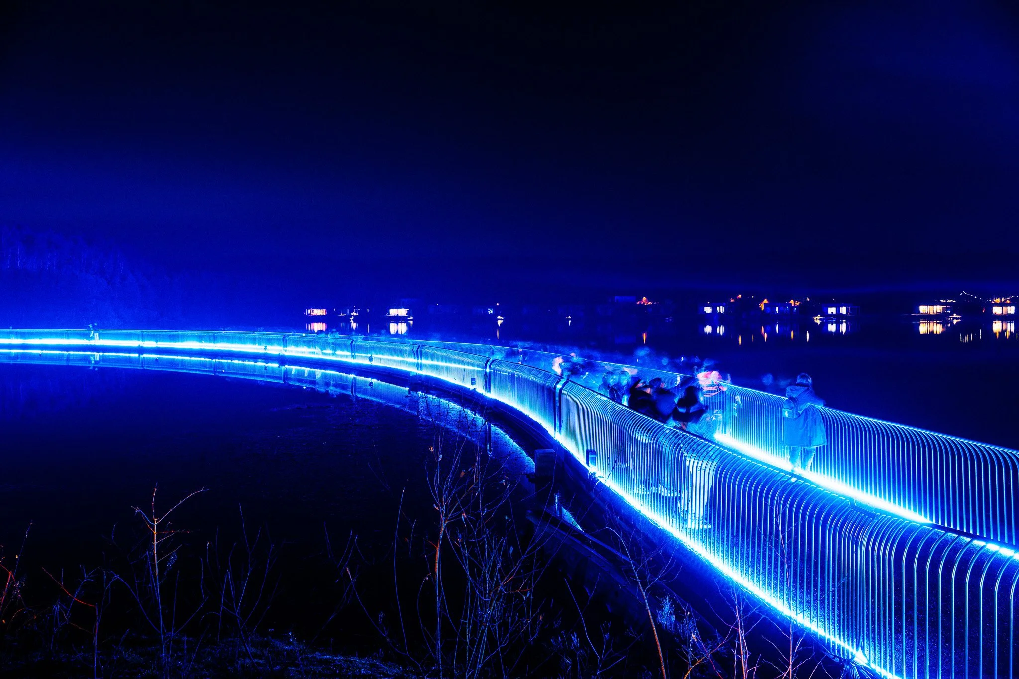Een verlichte wandelroute bij nacht met blauwe LED-verlichting langs een waterkant, mensen lopen langs de route onder een donkere hemel.