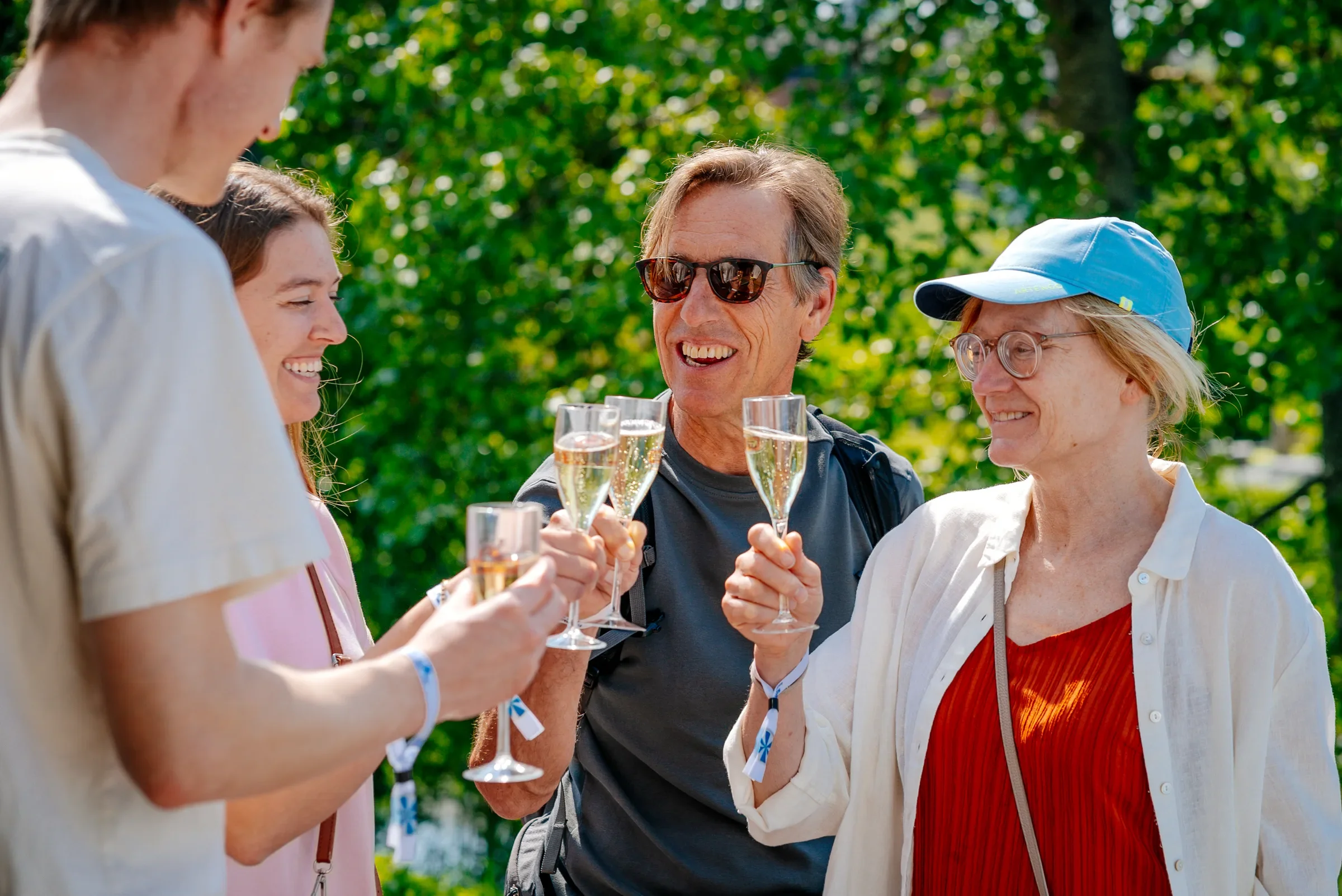 Vijf mensen tijdens een feestje in een park, ze proosten met glazen champagne en lachen, omgeven door groene bomen.