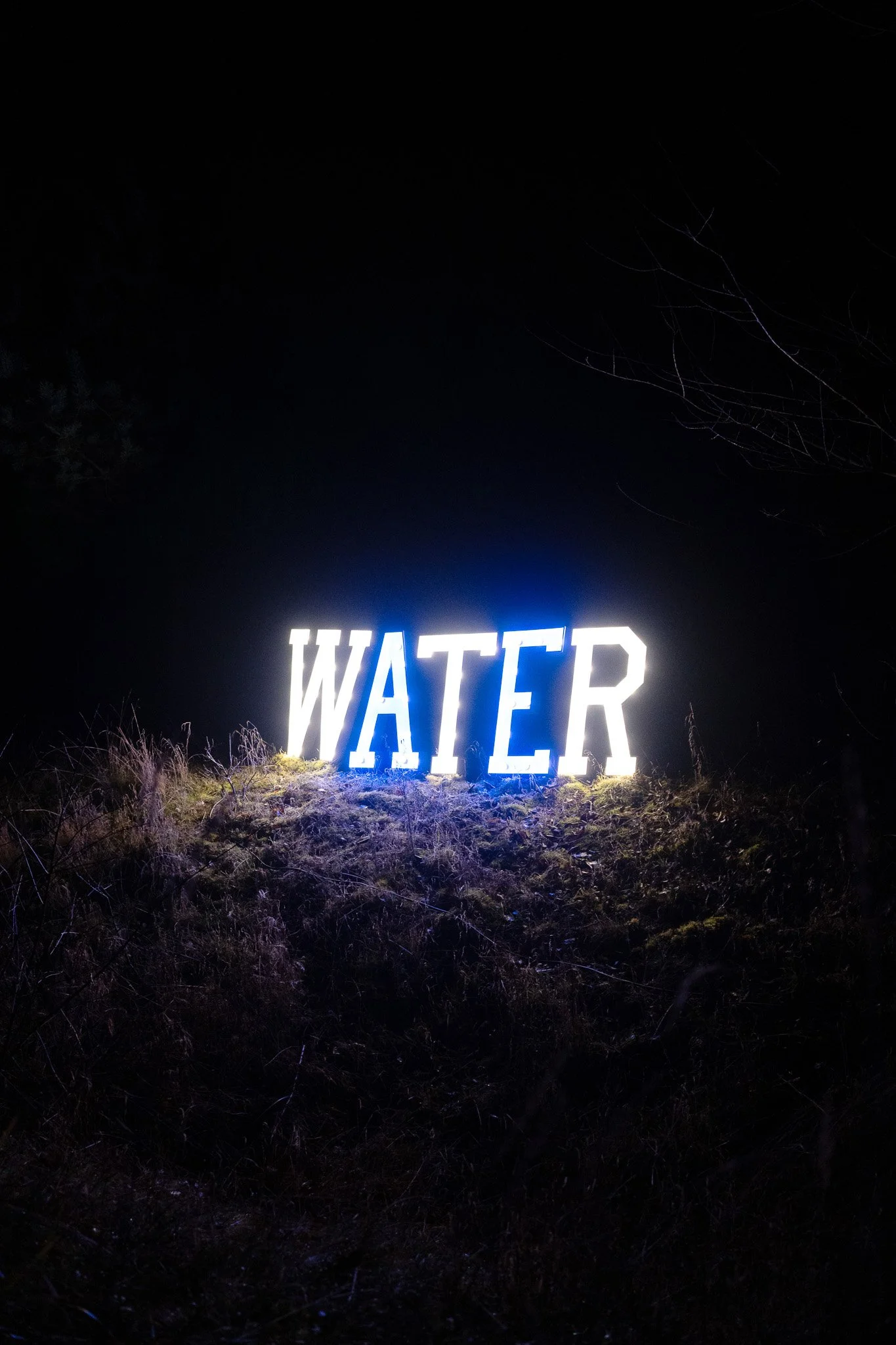 Verlichte tekst 'WATER' op een bord in een duistere omgeving, met een bosachtig achtergrond.