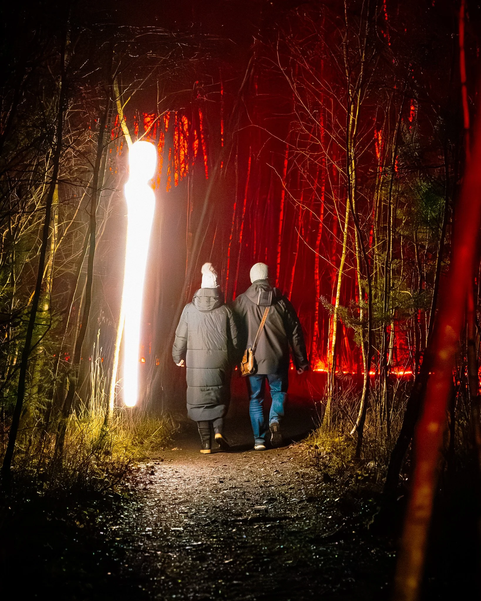 Twee mensen lopen 's avonds door een bos met verlichte paden en rode lichten, met lichtgevende objecten langs de route.