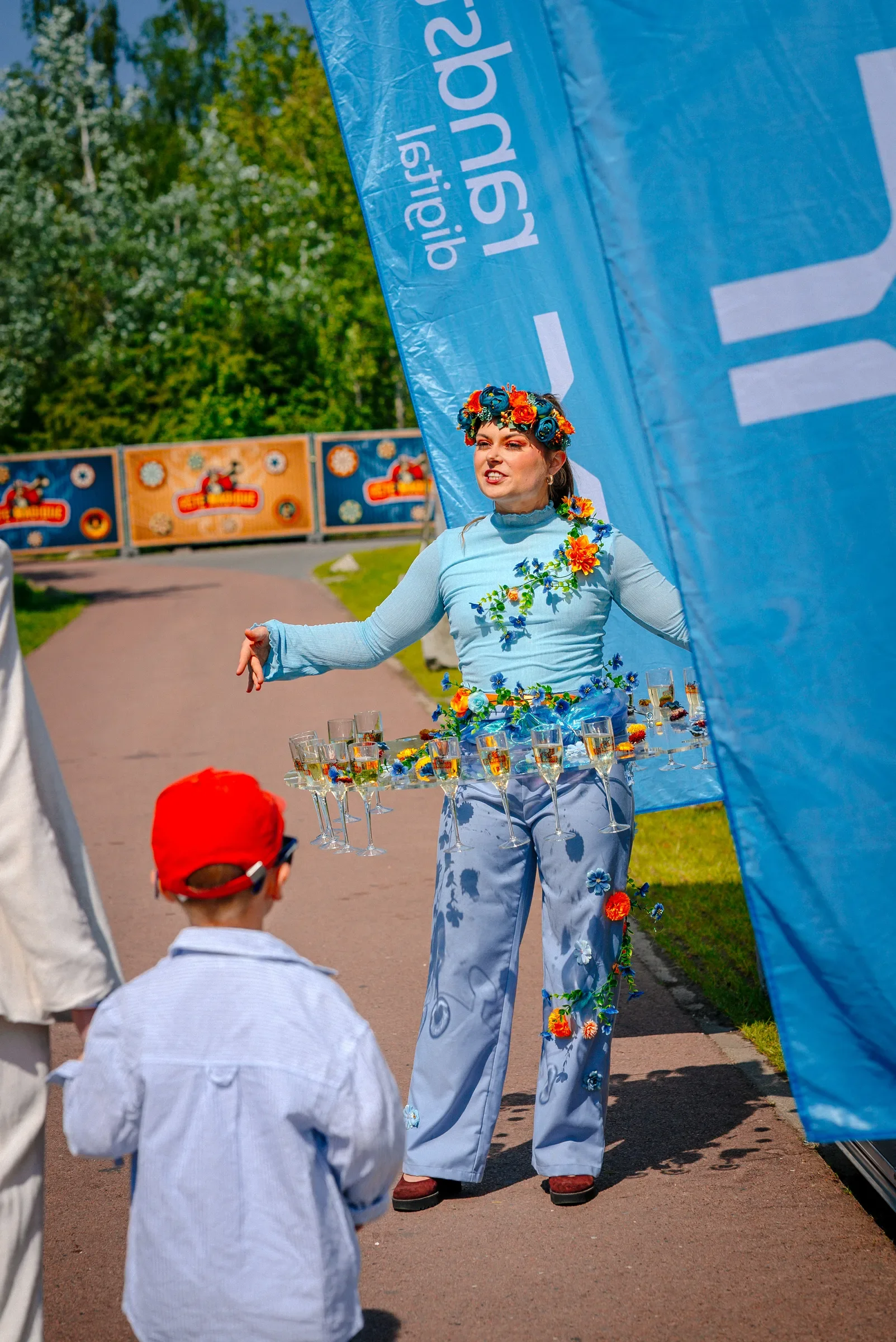 Vrouw in traditionele kleding met bloemen en wijn op een straat, met kinderen in de buurt, bij een blauw vlaggetje.