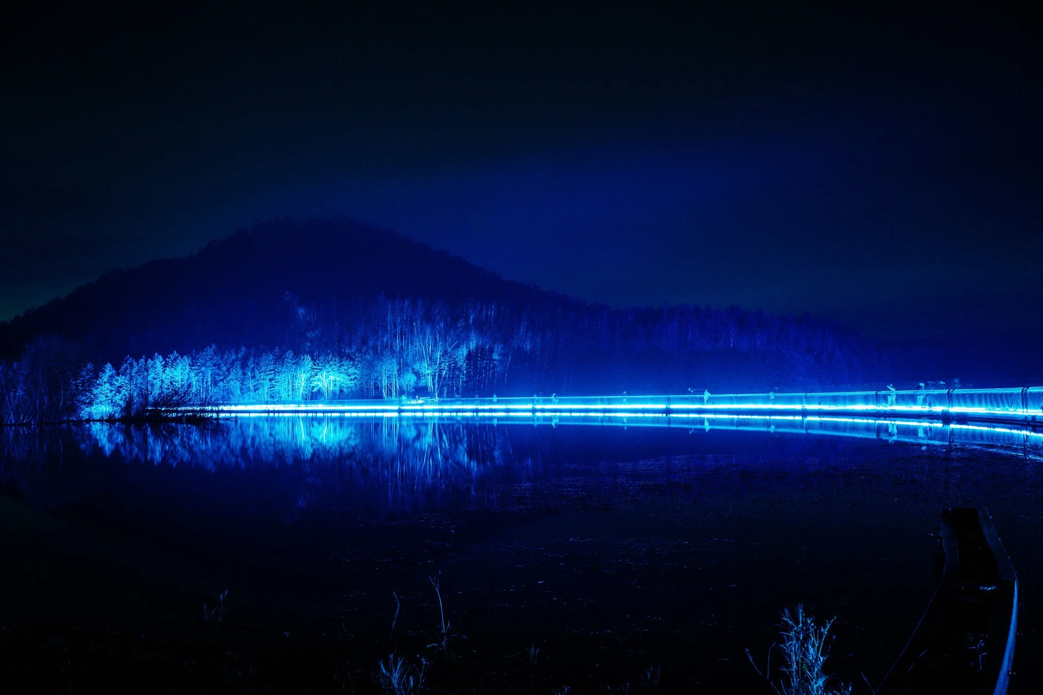 Een nachtlandschap met een berg en een bos, verlicht door blauwe lichten, langs een rustig meer dat de berg en bomen weerspiegelt.