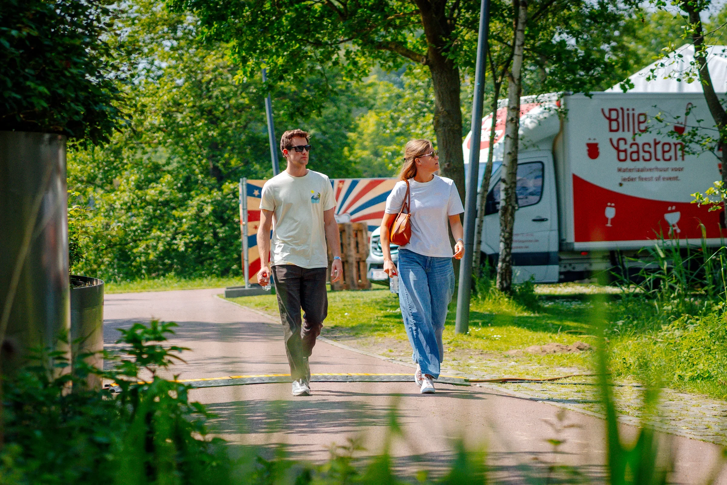 Twee mensen wandelen langs een pad in een park op een zonnige dag, met bomen en een voedseltruck op de achtergrond.