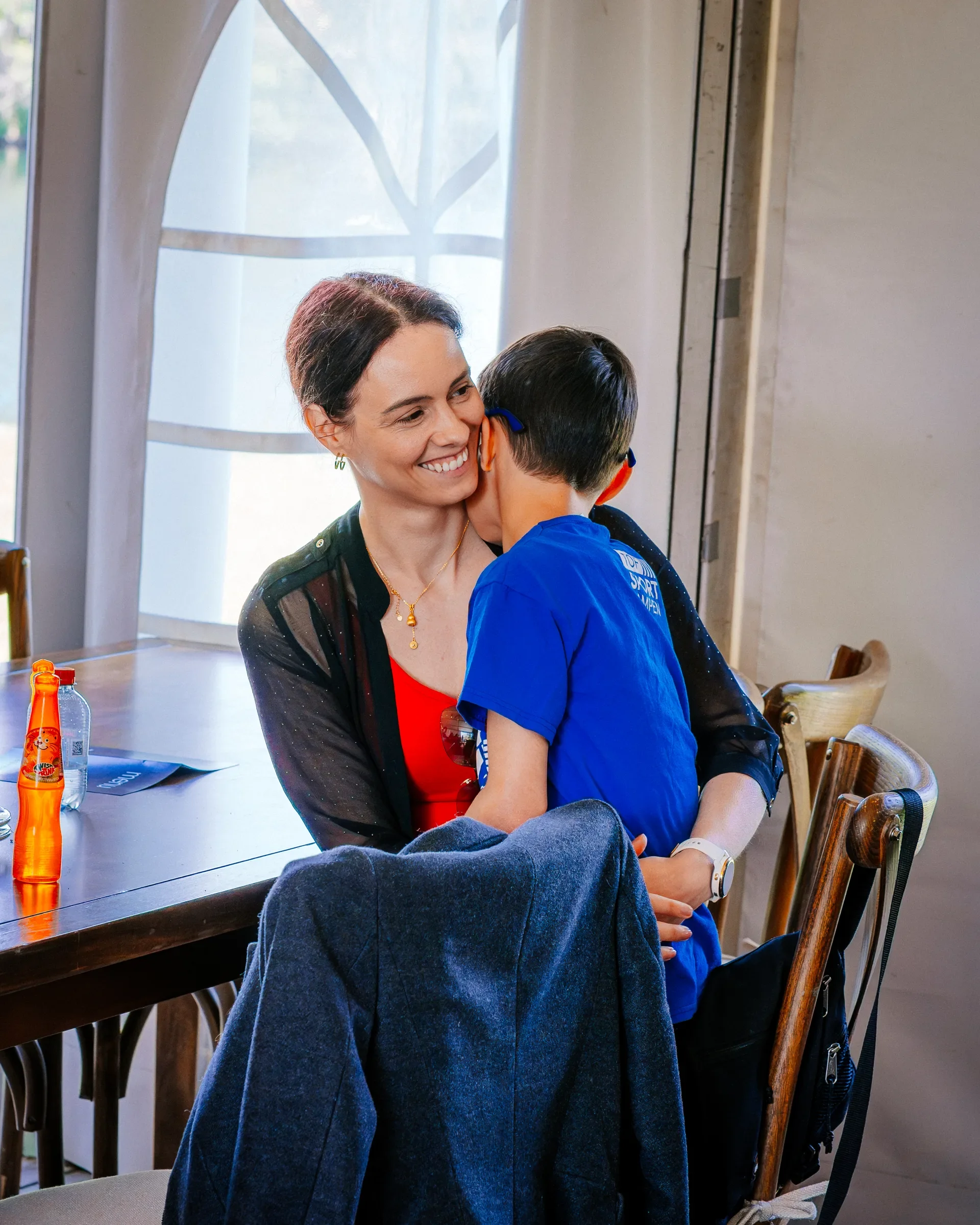 Een vrouw en een kind delen een knuffel in een restaurant of café. De vrouw glimlacht, het kind heeft de rug naar de camera.