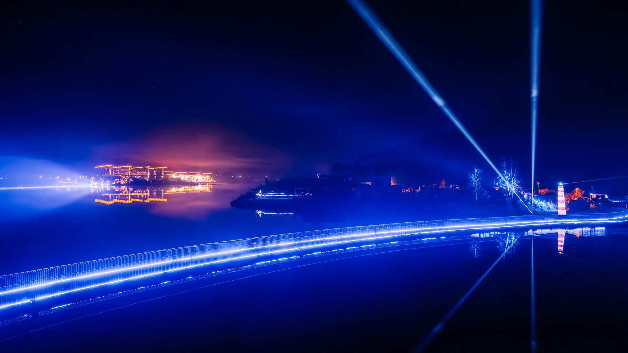 Lichtshow met blauwe laserstralen boven een watergebied, verlichte gebouwen en industrie in de achtergrond, reflecties op het water, 's nachts.