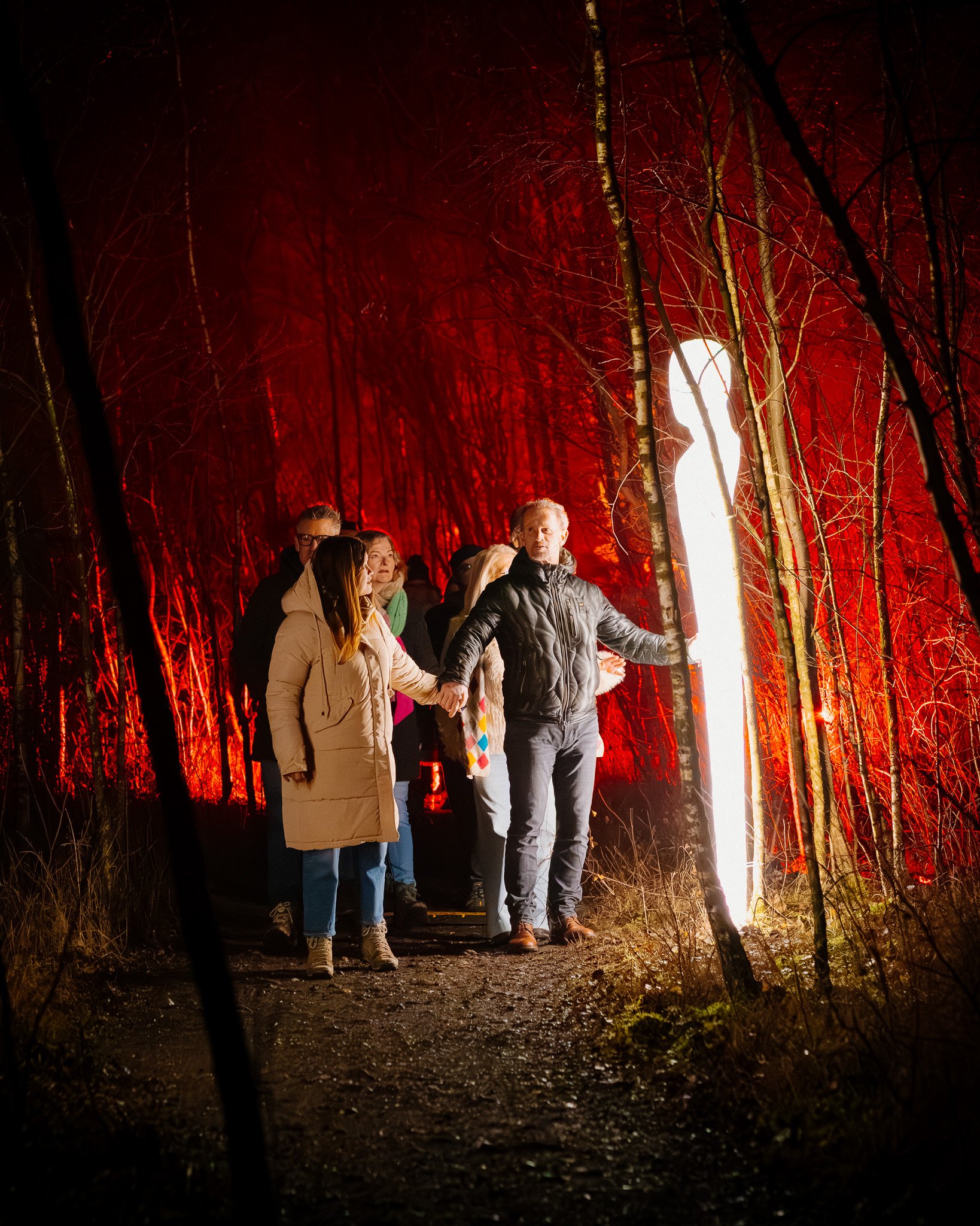 Een groep mensen houdt handen vast en wandelt door een donkere boslucht met Rode verlichting op de achtergrond en een intense lichtstraal die van achter licht geeft.
