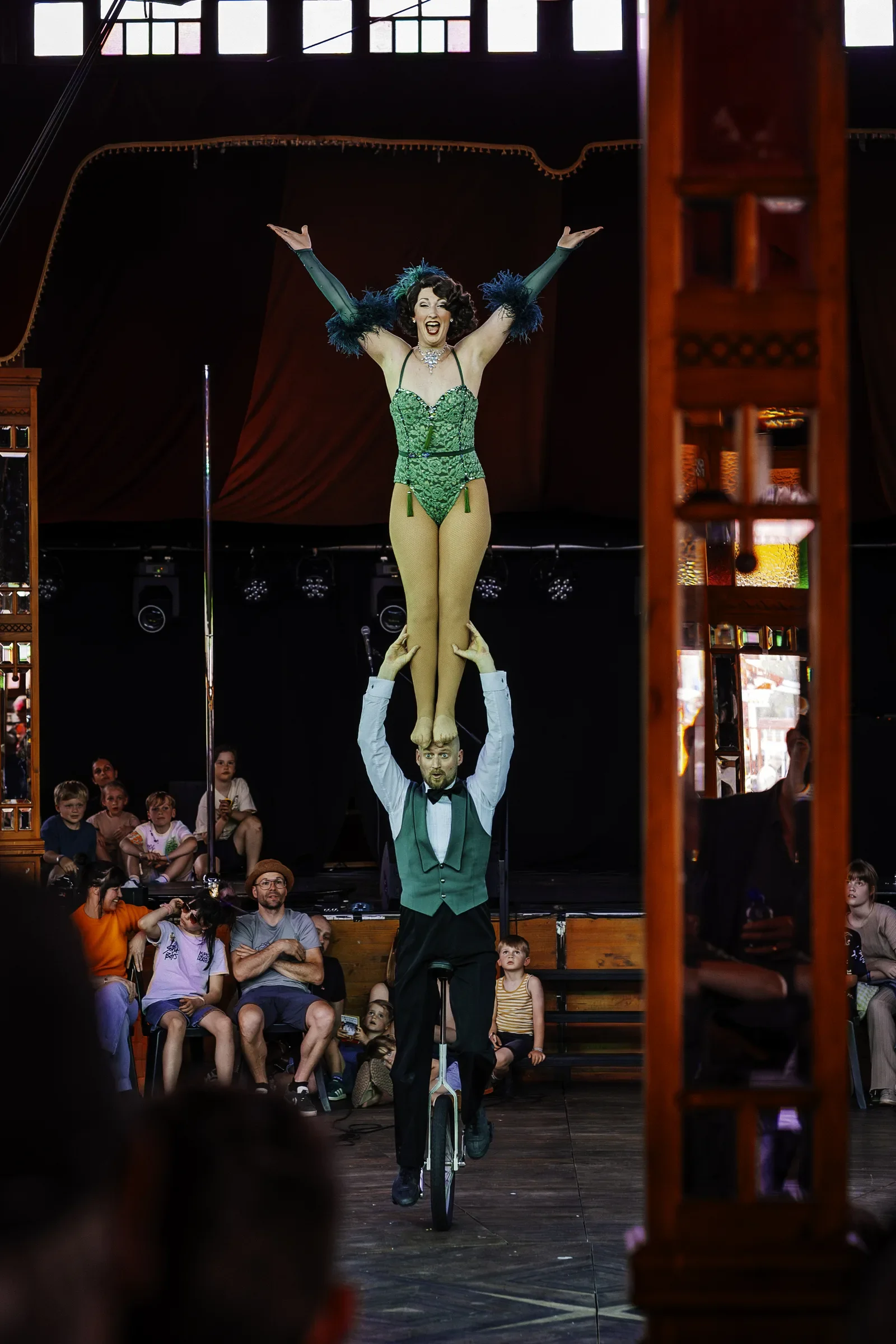 Eencircusact met een vrouw in een groene kostuum die op een eenwieler rijdt, terwijl een man haar ondersteunt en haar optilt met zijn handen. Er zitten mensen in het publiek die kijken naar de show.
