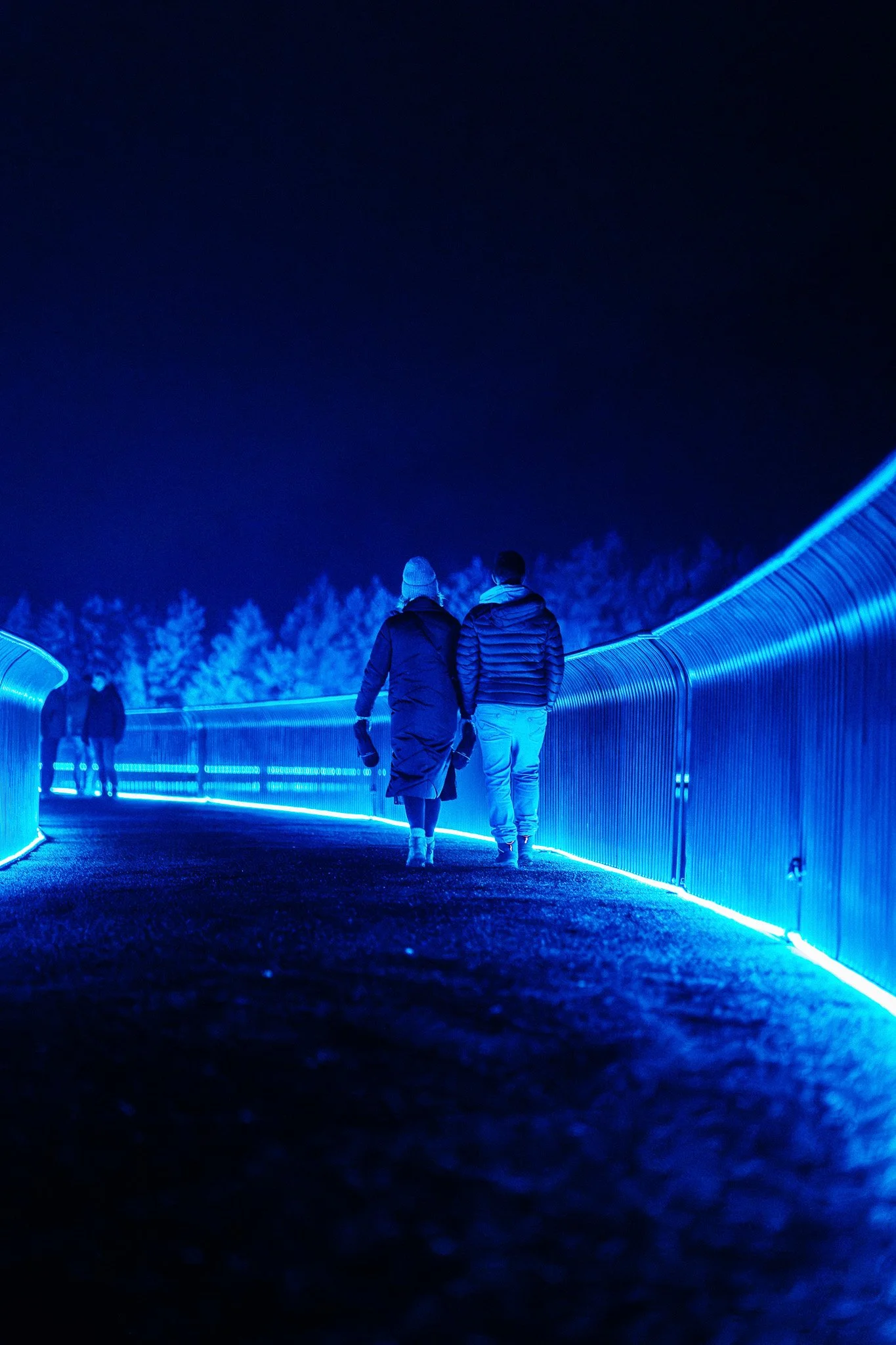 Twee mensen lopen over een met blauwe neonlichten verlichte voetgangersbrug 's nachts, met een groep mensen op de achtergrond.