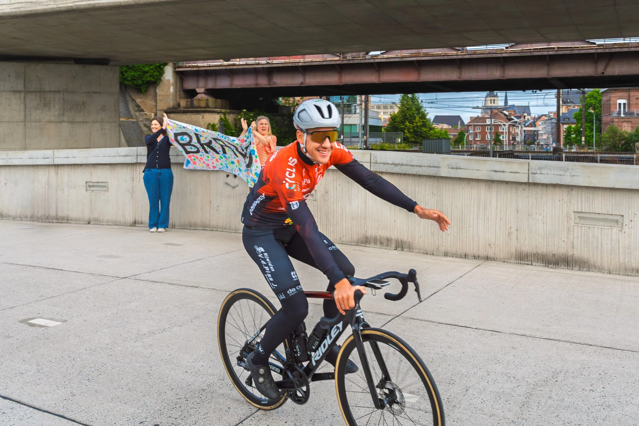 Cyclis renner fietst lachend langs supporters onder een brug