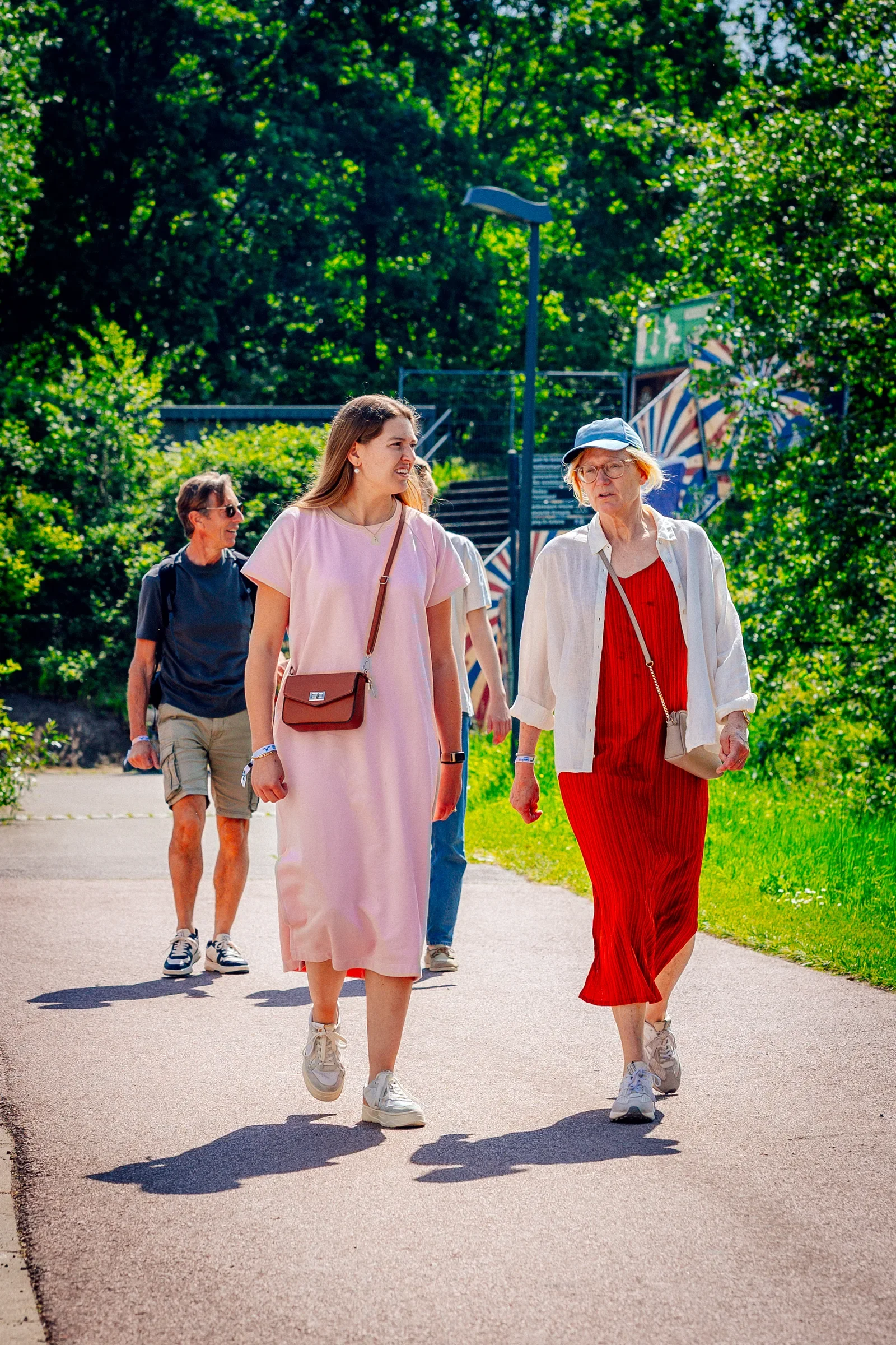 Vijf vrouwen wandelen op een pad in een park, omgeven door groene bomen, zomers gekleed en genieten van het buitenseizoen.