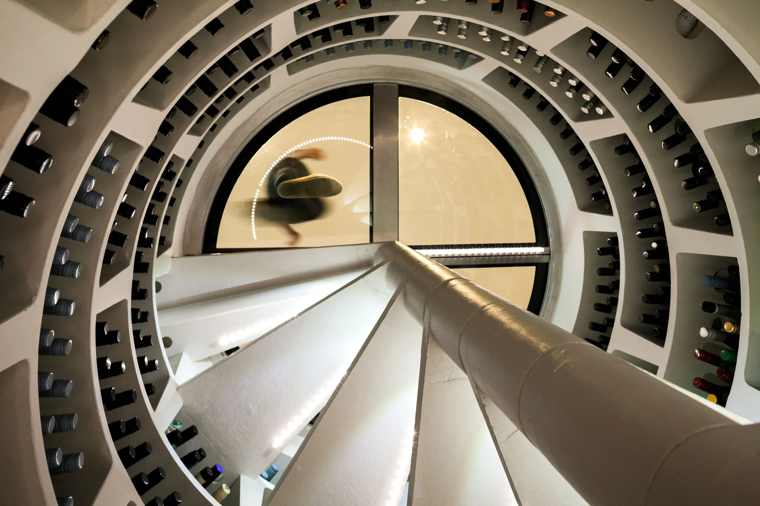 View through a circular wine bottle storage with bottles placed on multiple concentric shelves, and a person walking past the glass door at the bottom of the storage area.