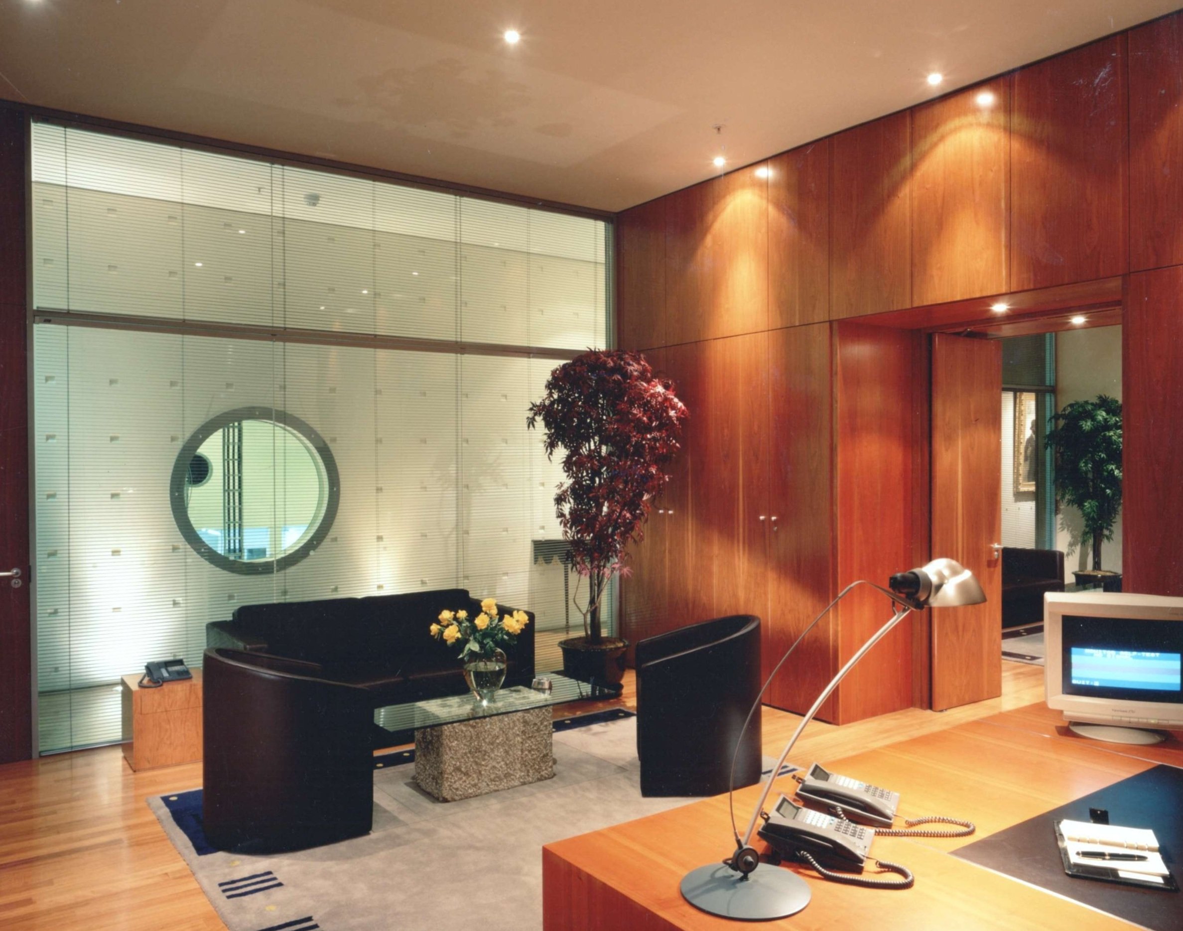 An indoor office or lobby area with wooden walls, large window blinds, a small glass table with a flower arrangement, and a few chairs. There are also some electronic devices and lamps.