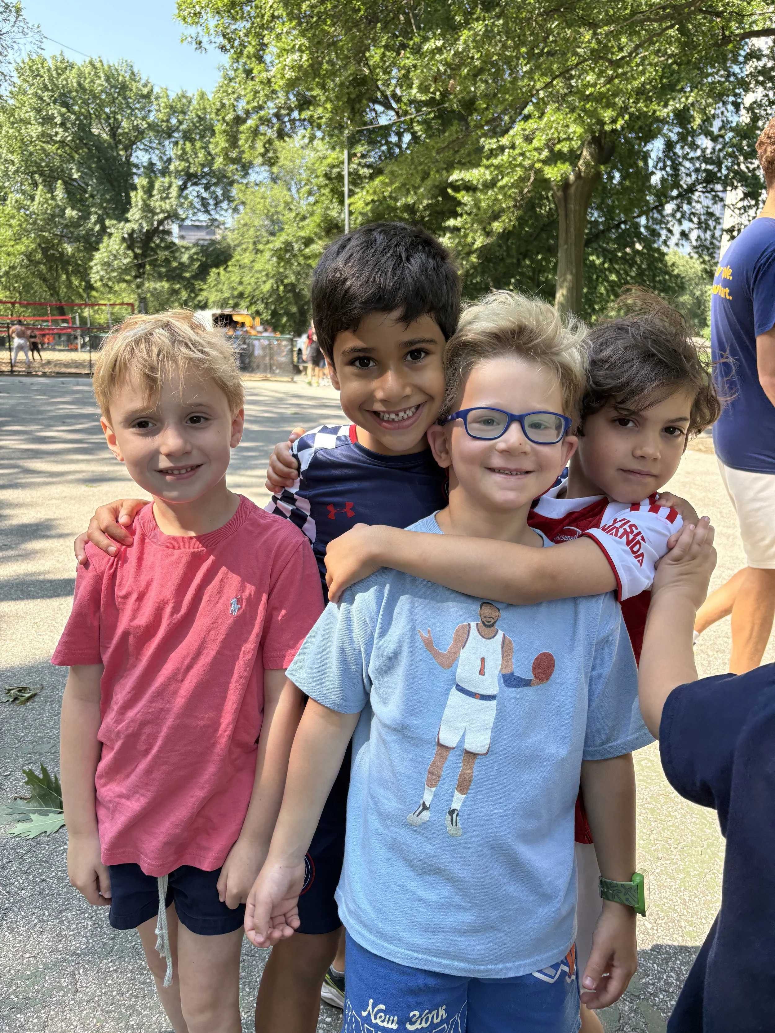 Four children smiling and hugging outdoors on a sunny day, with trees and a playground in the background.