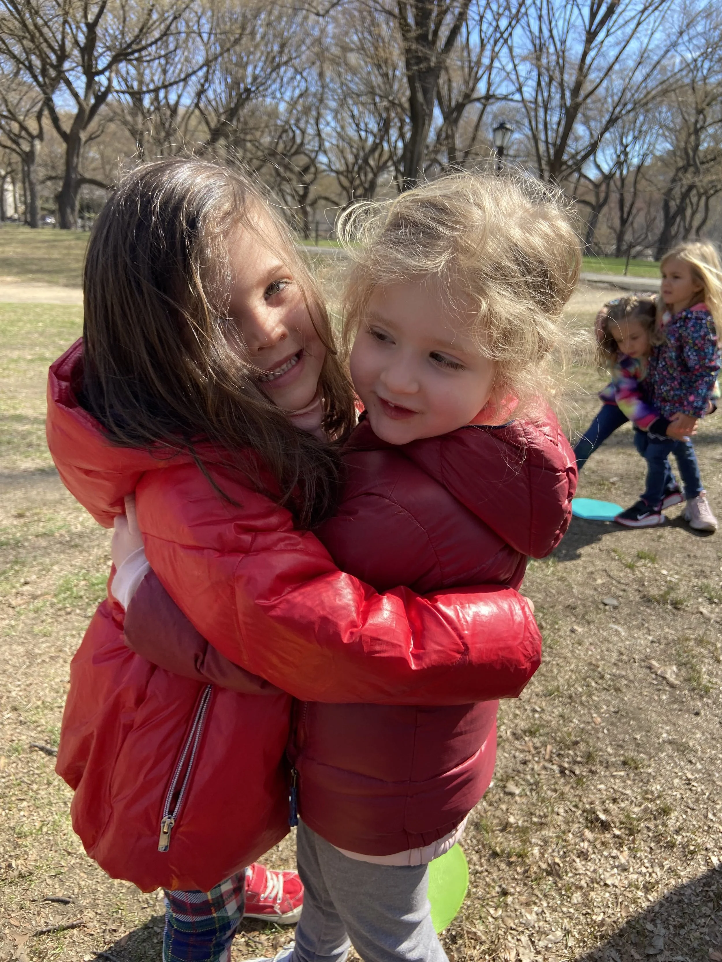 Two young girls hugging outdoors in a park on a sunny day, with a few other children playing in the background.