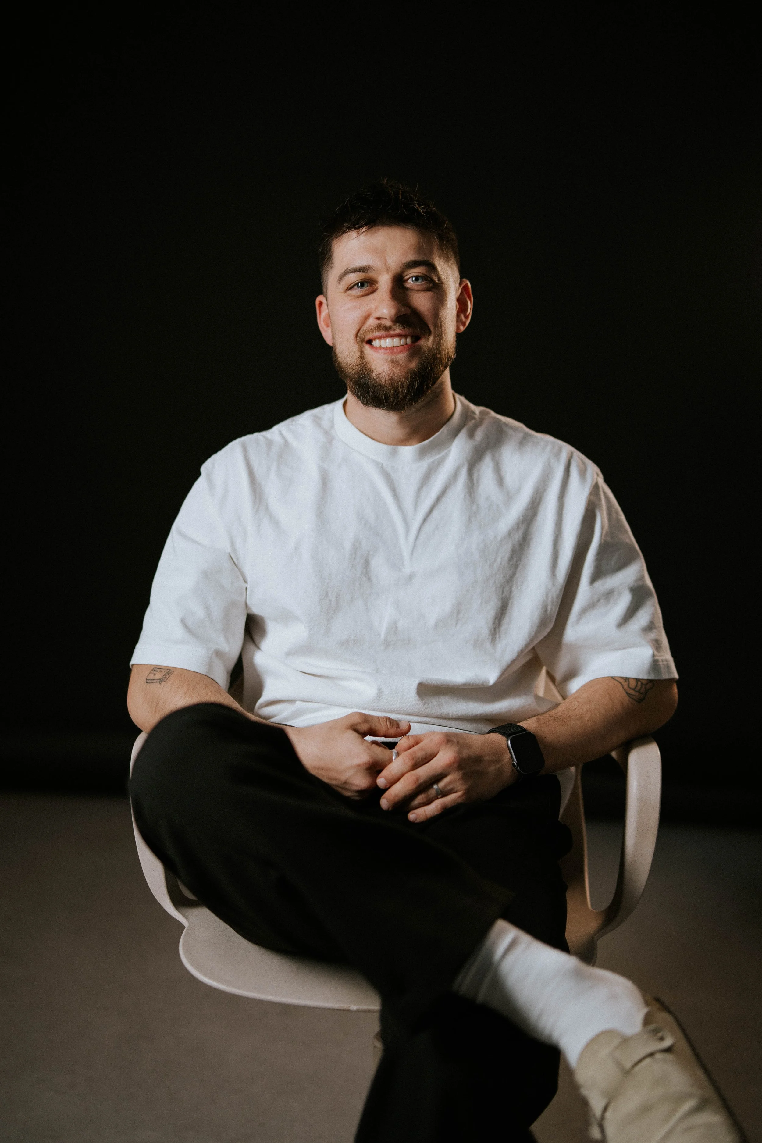 Young man with a beard smiling, sitting with legs crossed, wearing a white t-shirt, black pants, white sneakers, and a smartwatch, against a dark background.