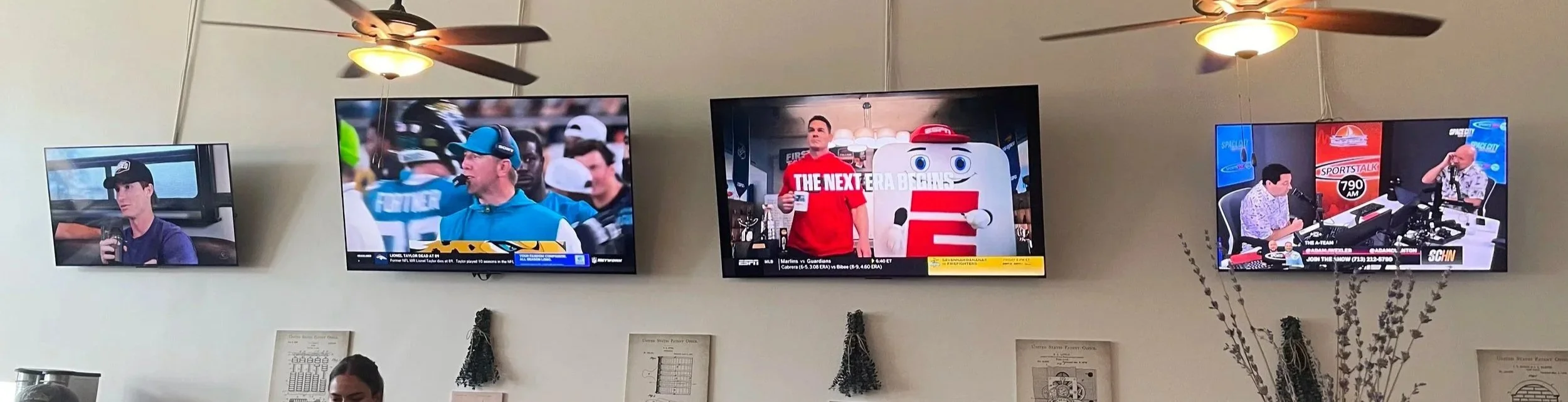 Multiple flat-screen TVs mounted on a wall, showing different sports and entertainment programs in a room with ceiling fans and hanging decorative plants.