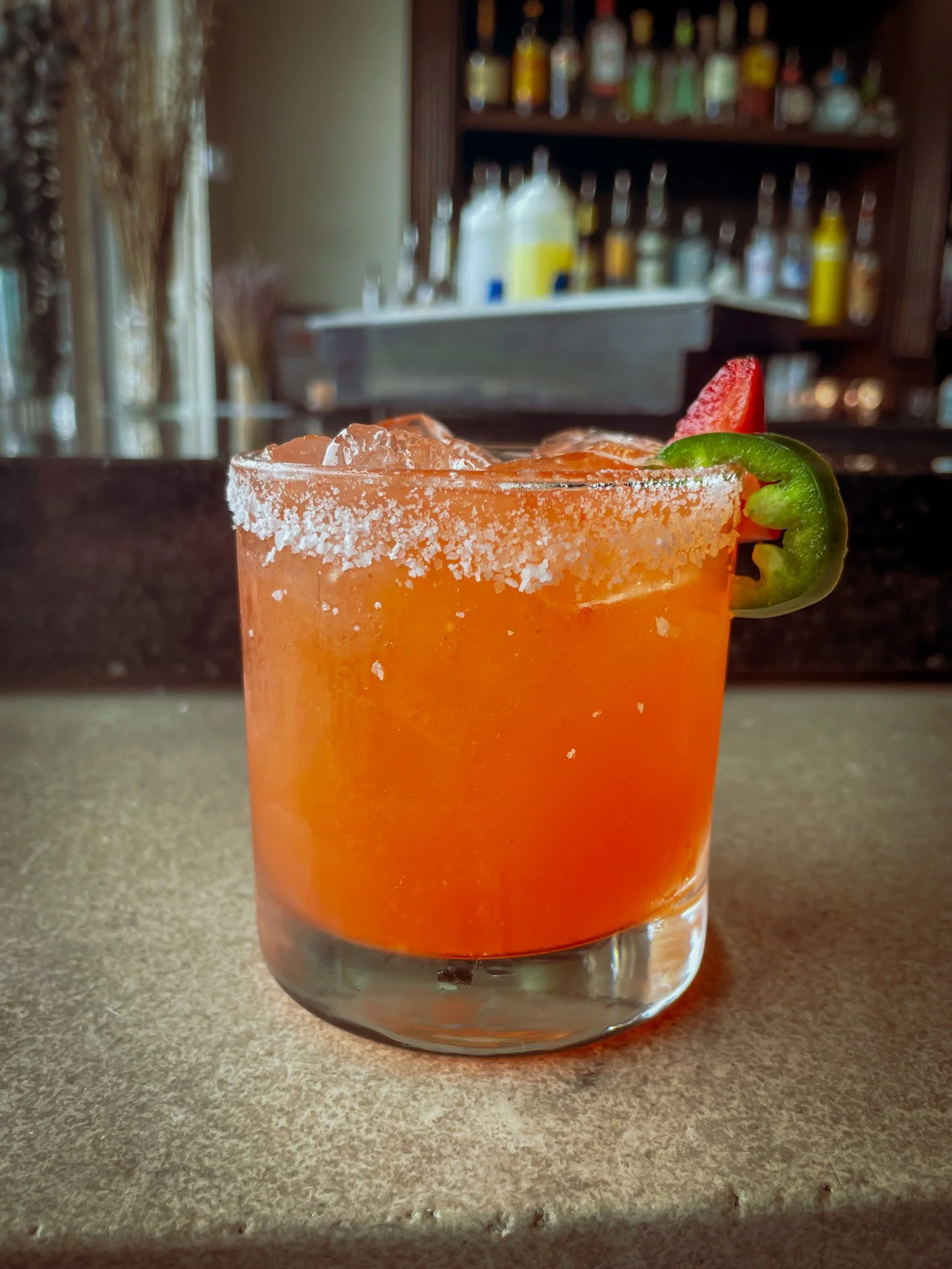 A cocktail in a short glass with an orange hue, garnished with a slice of green bell pepper and a strawberry, with a salted rim.