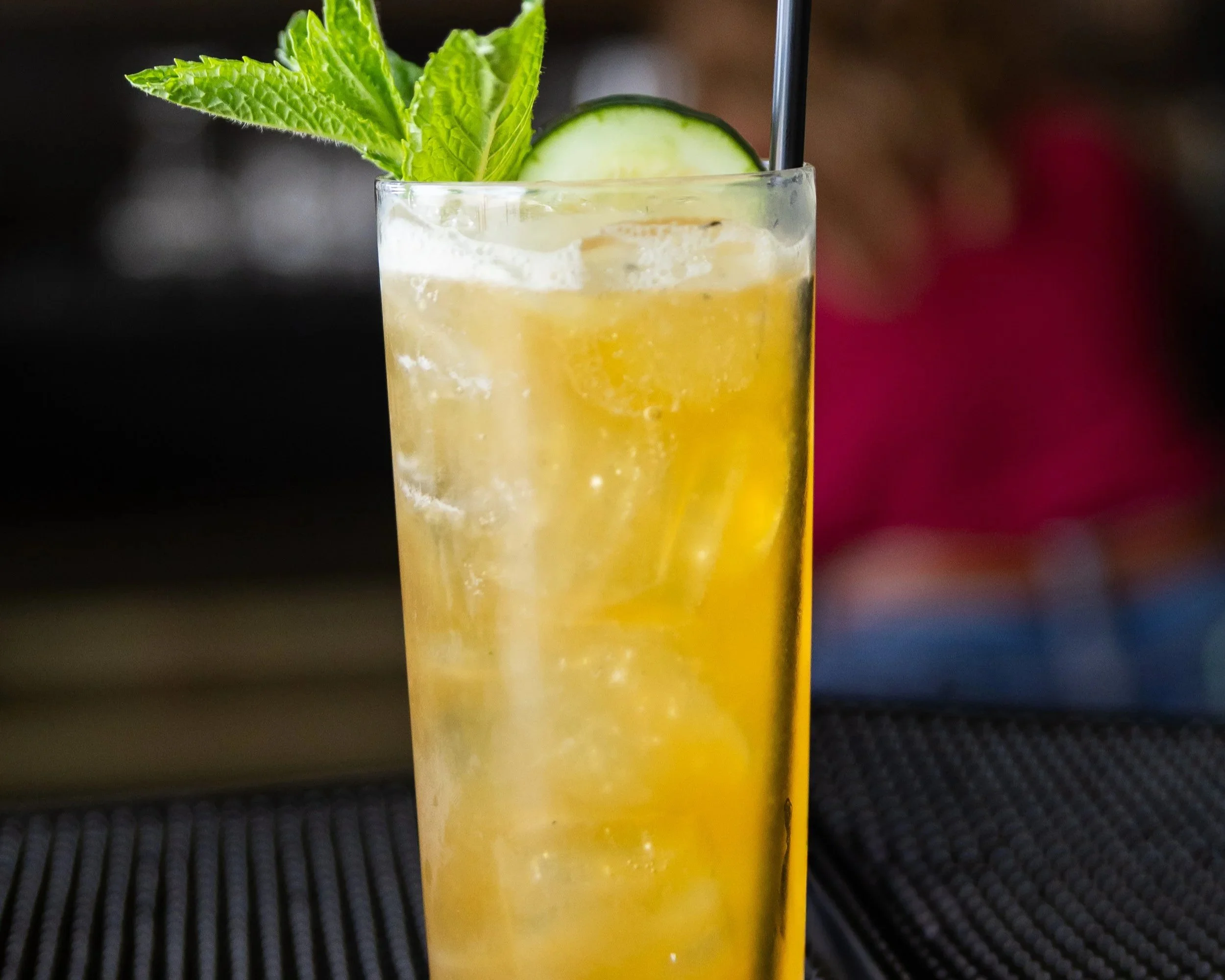 A tall glass of yellow cocktail with ice, garnished with a cucumber slice and fresh mint leaves, with a straw inside.
