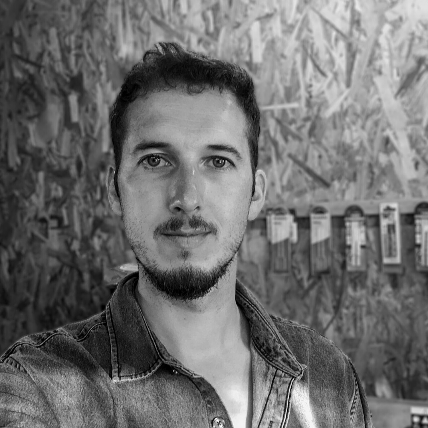 Black and white portrait of a man wearing a denim shirt in front of a textured wall with tools hanging in the background.