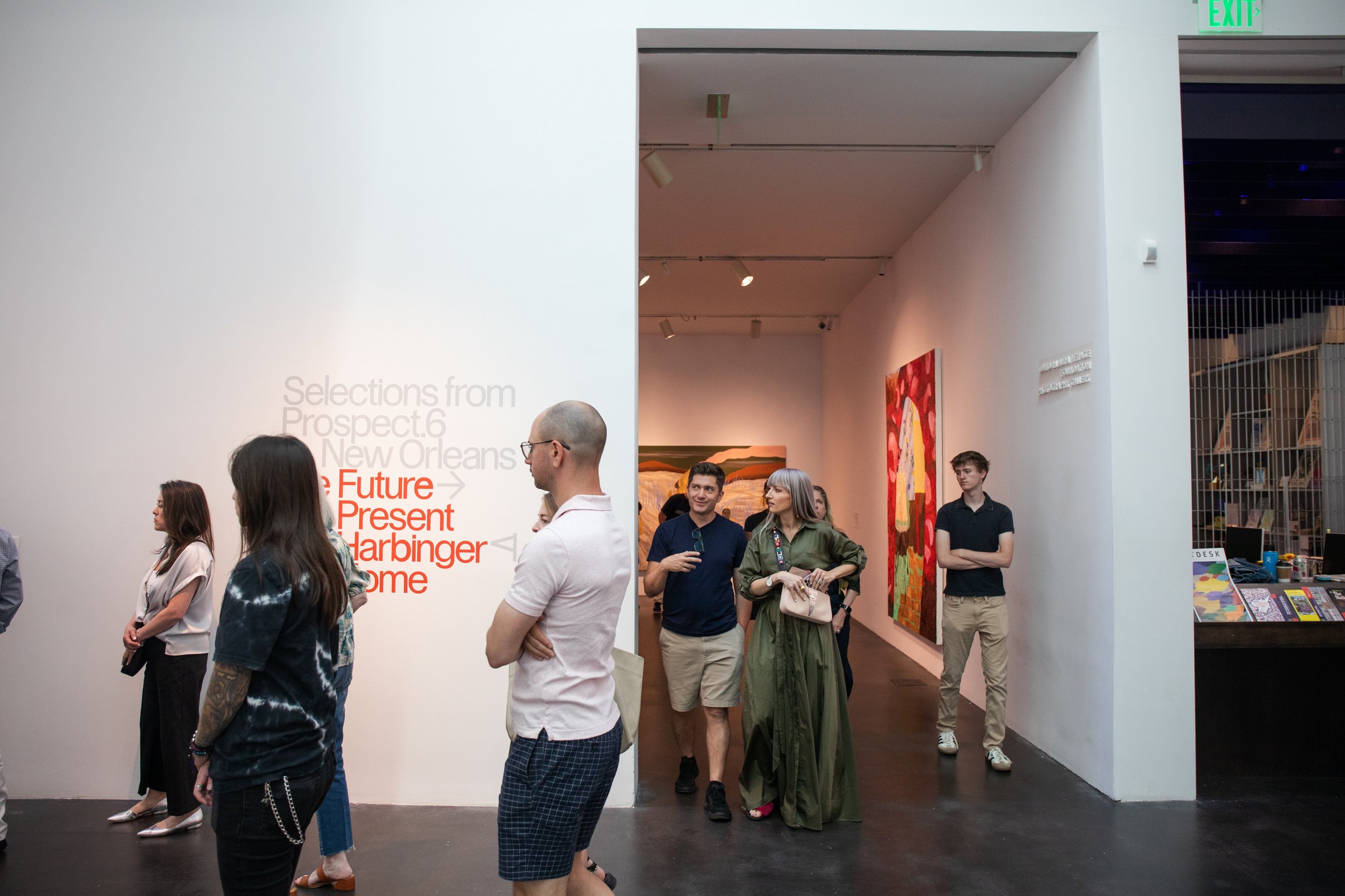 Biennial Culture Club at MCA. People viewing art in a gallery with one artwork featuring a large colorful abstract painting. A white wall has text with red and gray fonts about art selections, and there is a reception desk with books and a computer.