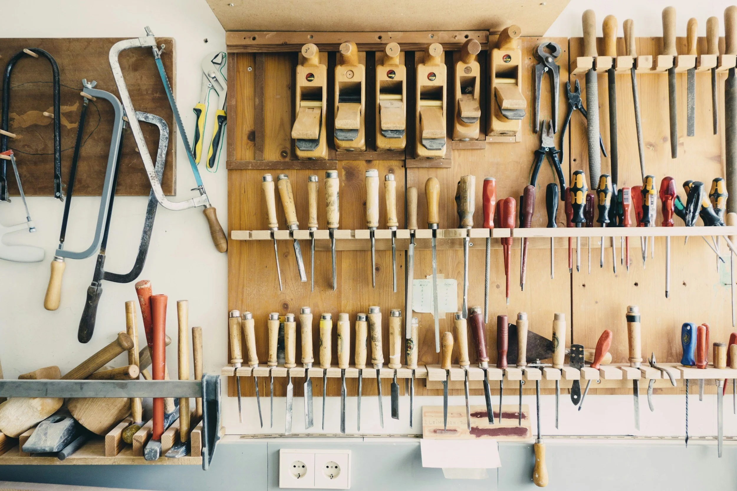 Woodworking tools organized on a workshop wall, including saws, chisels, planes, pliers, screwdrivers, and files.