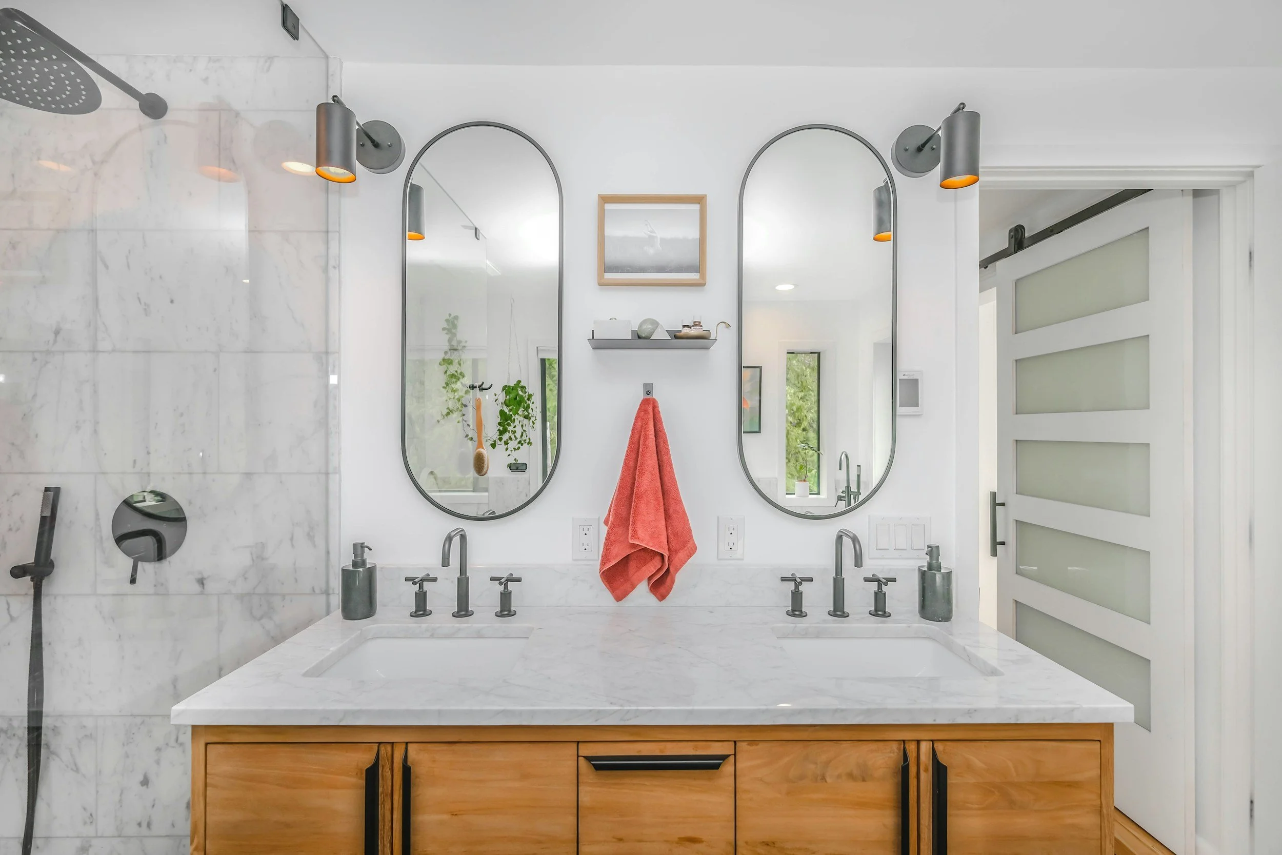 Modern bathroom with double sink vanity, oval mirrors, marble countertop, wooden cabinets, and a glass shower enclosure.