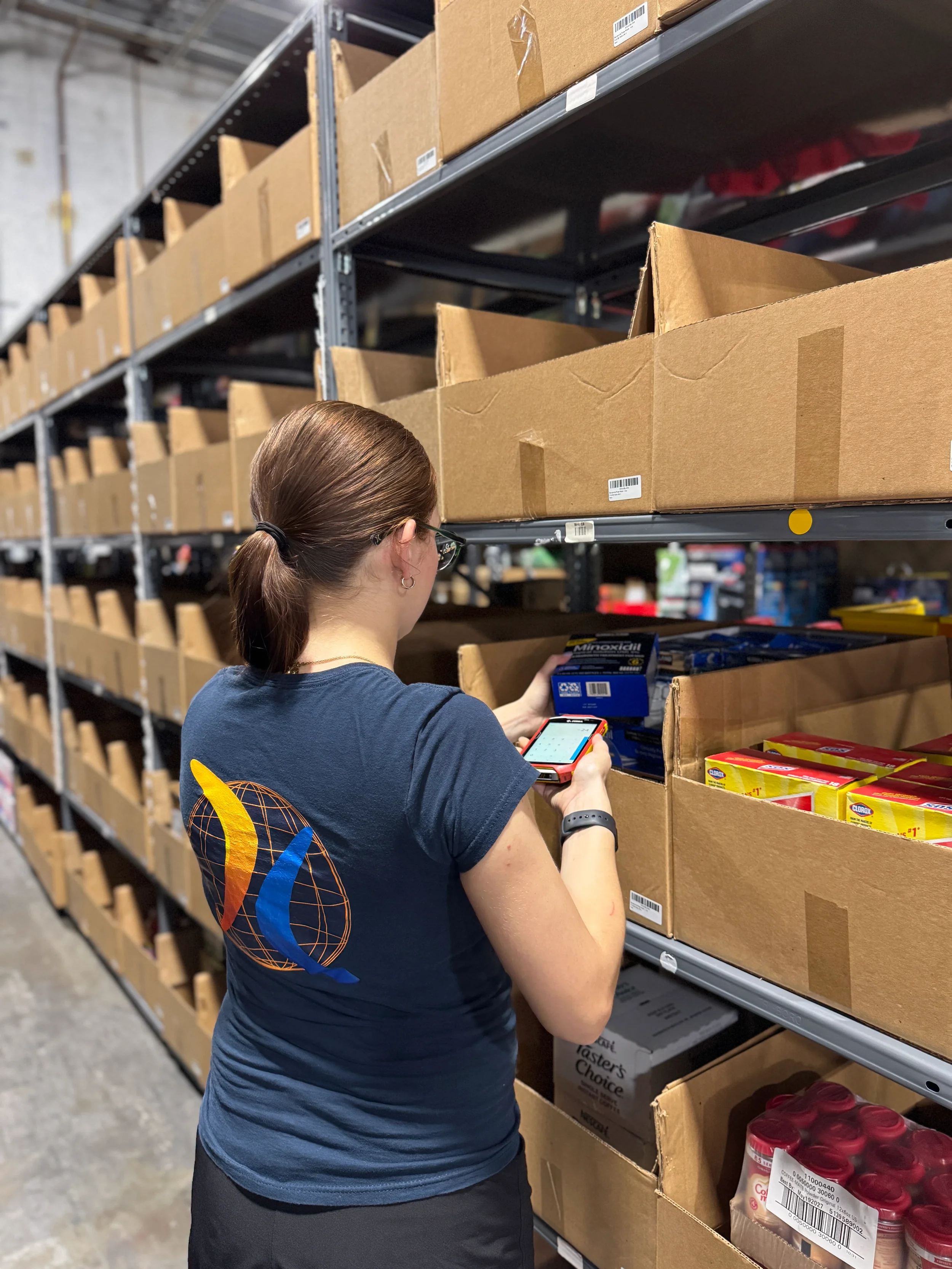 A woman with brown hair tied back, wearing glasses and a dark blue T-shirt with an orange and yellow globe logo on the back, is looking at a handheld electronic device while shopping in a store aisle with cardboard shelves stocked with various boxed products.