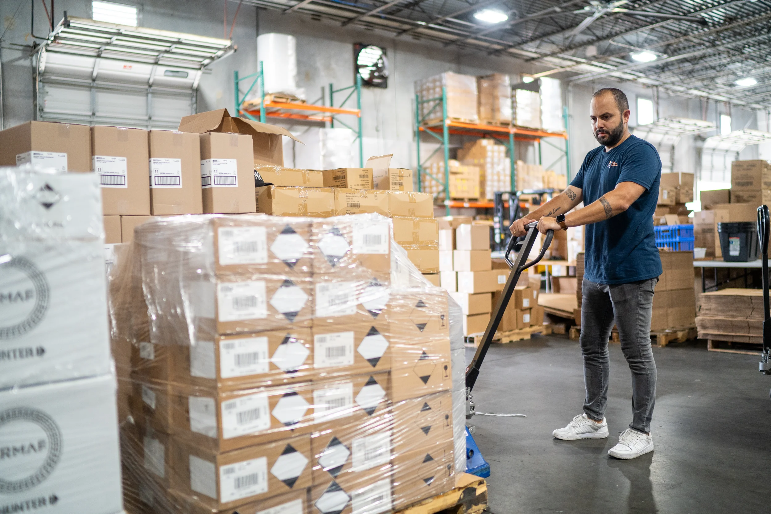 A man operates a pallet jack in a warehouse filled with stacked boxes and pallets.