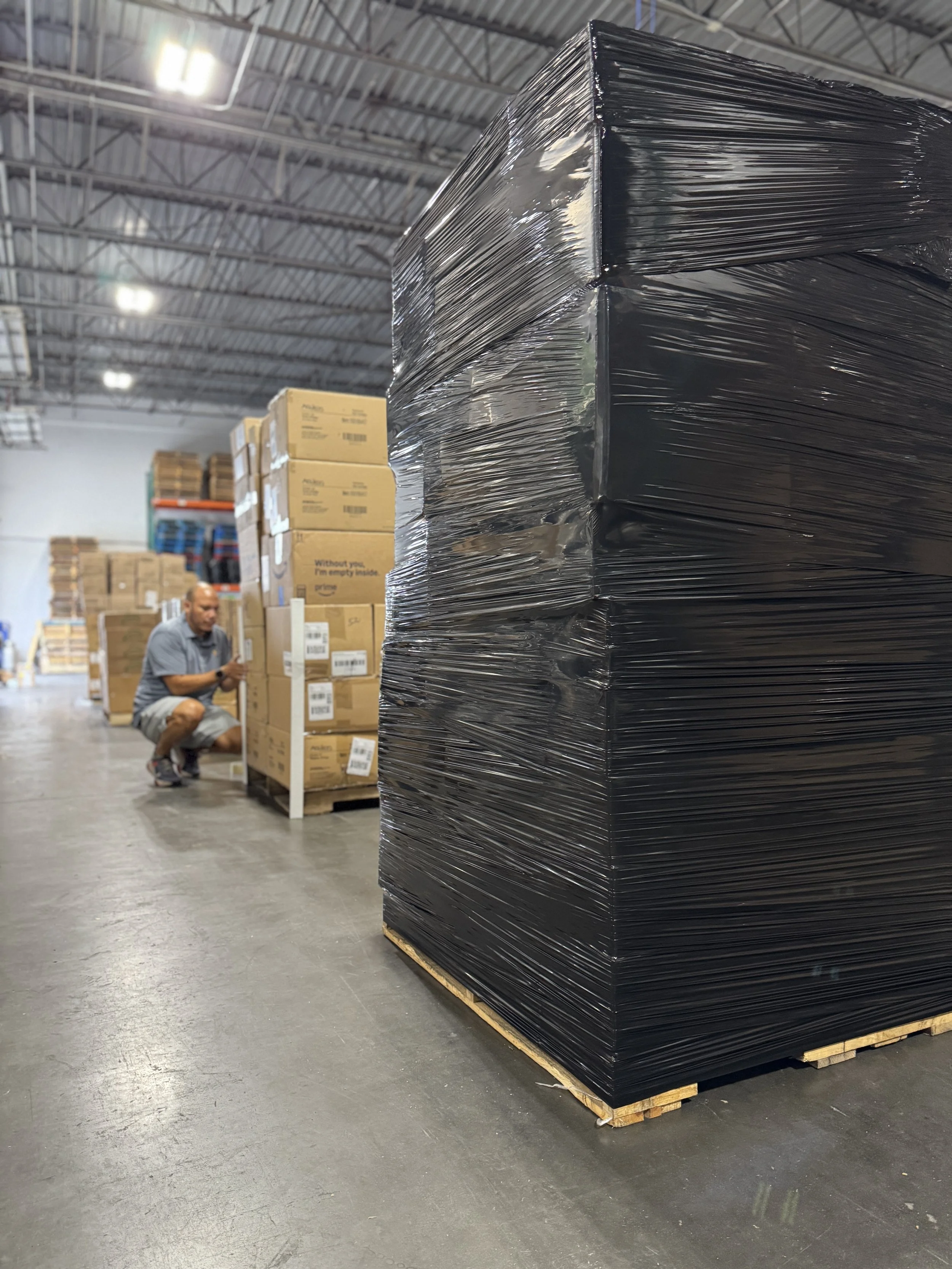 A warehouse with pallets of stacked boxes and a man squatting and looking at his phone.