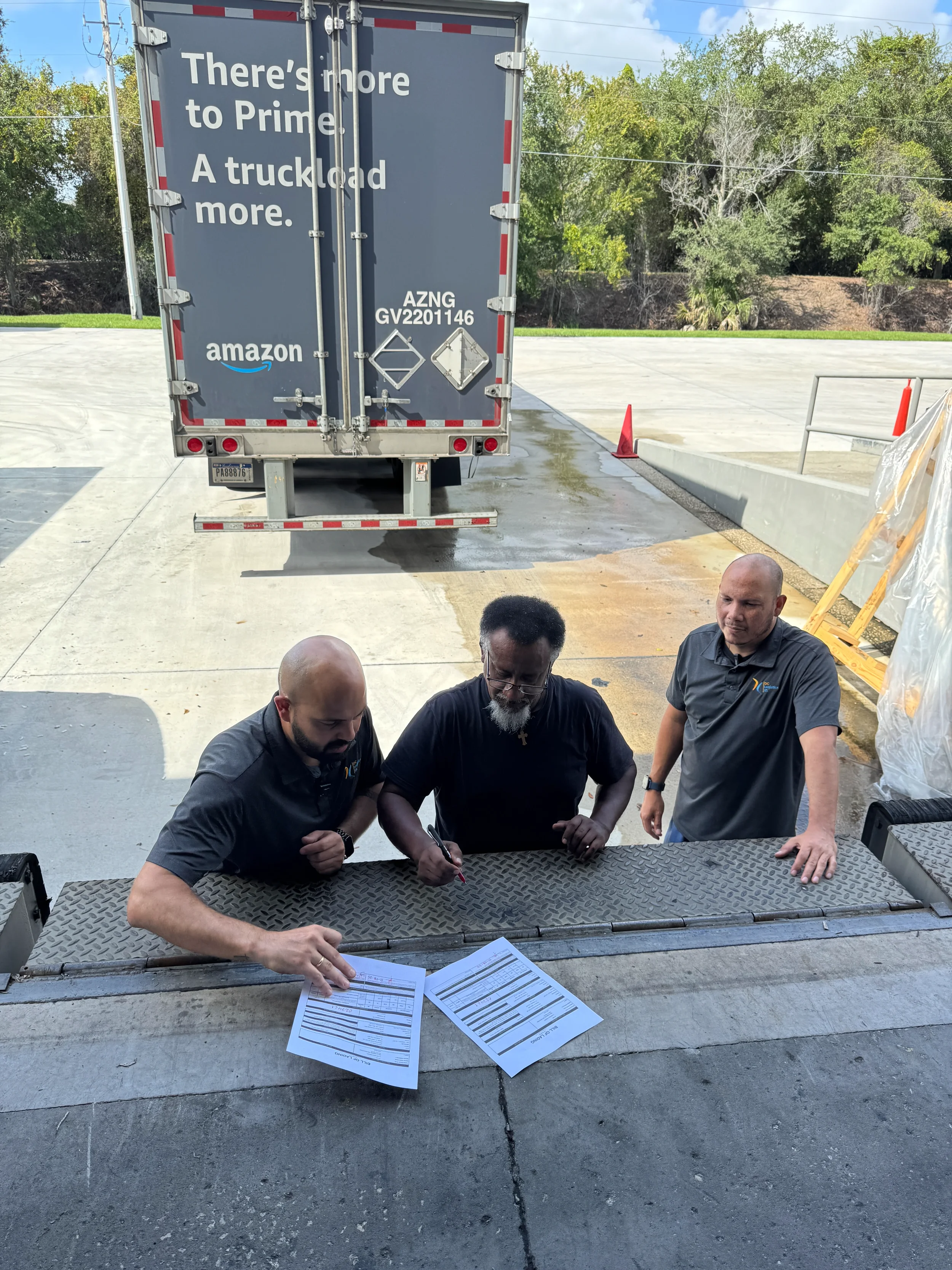 Three men are reviewing documents while standing at a loading dock, with a truck in the background and a parking lot and trees behind them.