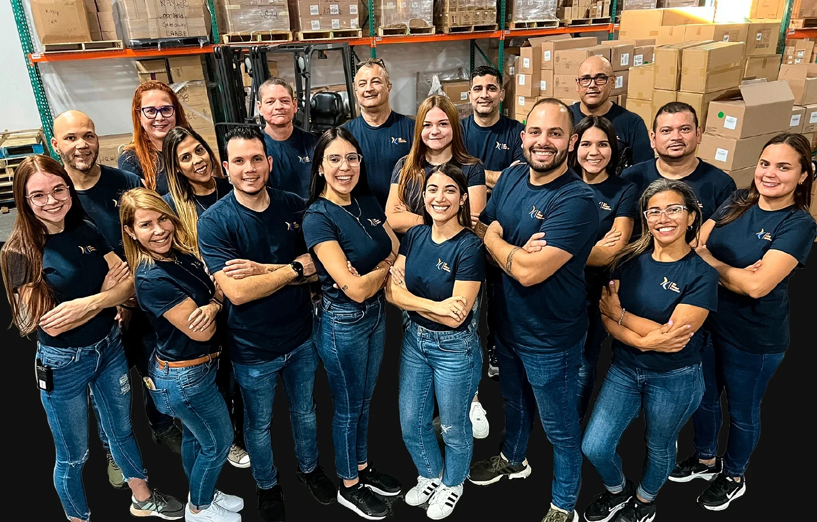 Group of 17 people standing in a warehouse, wearing matching blue shirts with a logo, smiling at the camera.