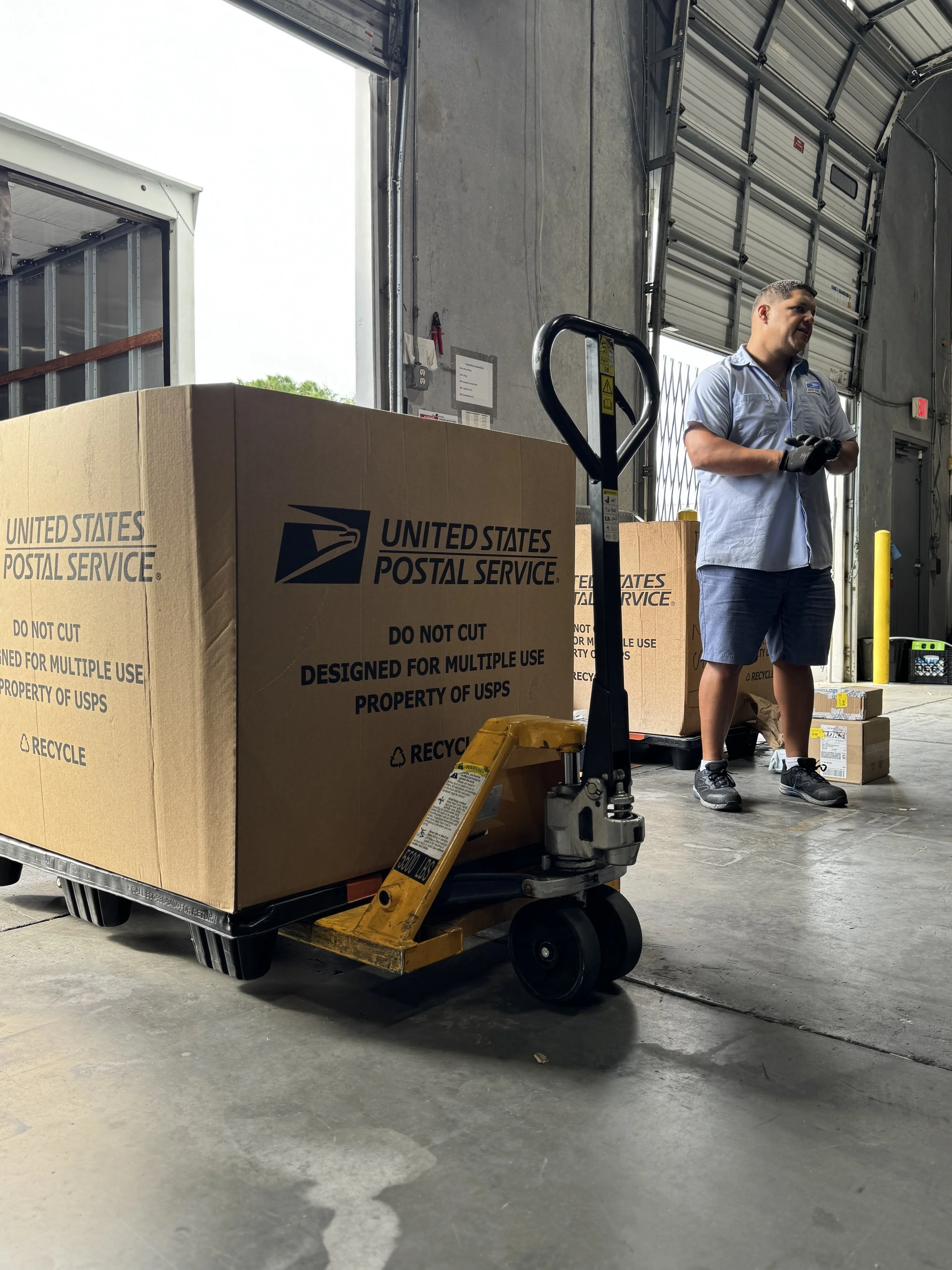 A man standing inside a warehouse or distribution center near a pallet of large USPS boxes and a pallet jack, with an open garage door in the background.