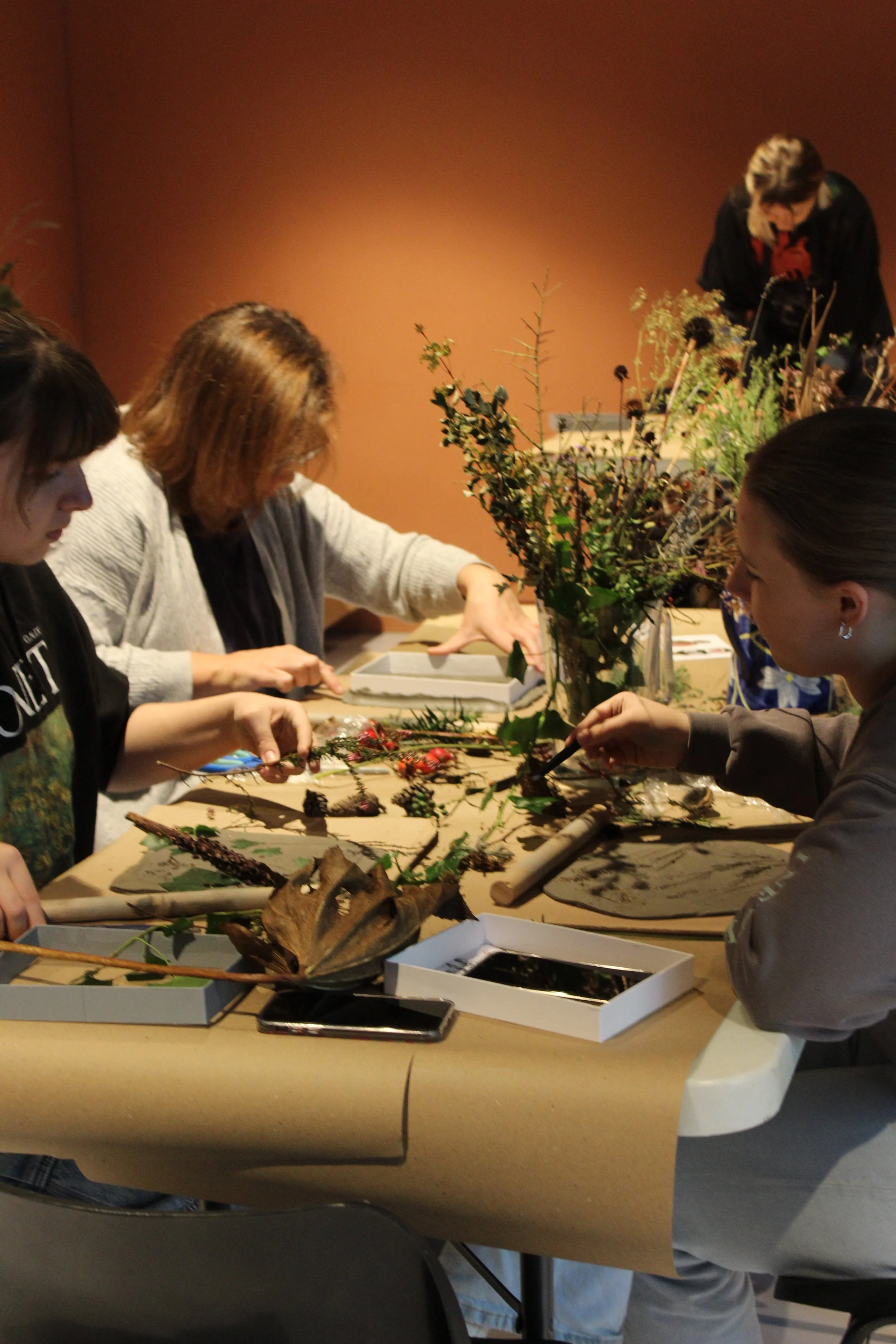 People participating in Eloise Pilbeam's Botanical Plaster Casting Workshop. Woman are sitting at a table with flowers, leaves, and natural objects, engaging in a craft or floral arranging activity.