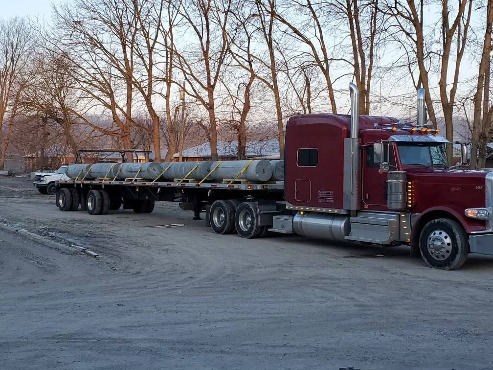 Red semi-truck hauling metal pipes on a flatbed trailer on a dirt road with leafless trees in the background.