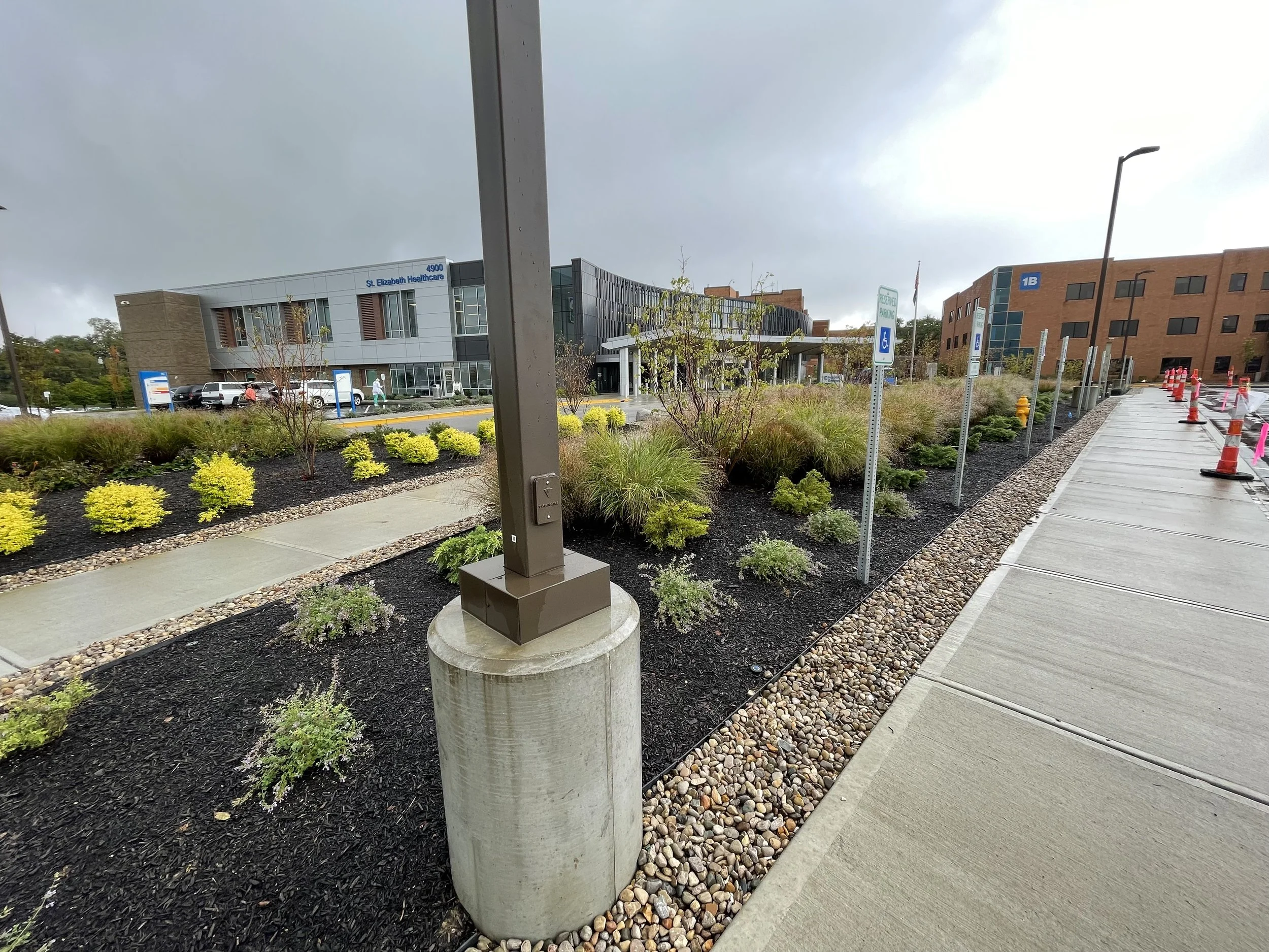 View of a hospital entrance with landscaped bushes and trees, parking lot, and a sidewalk with orange cones alongside. Overcast sky.