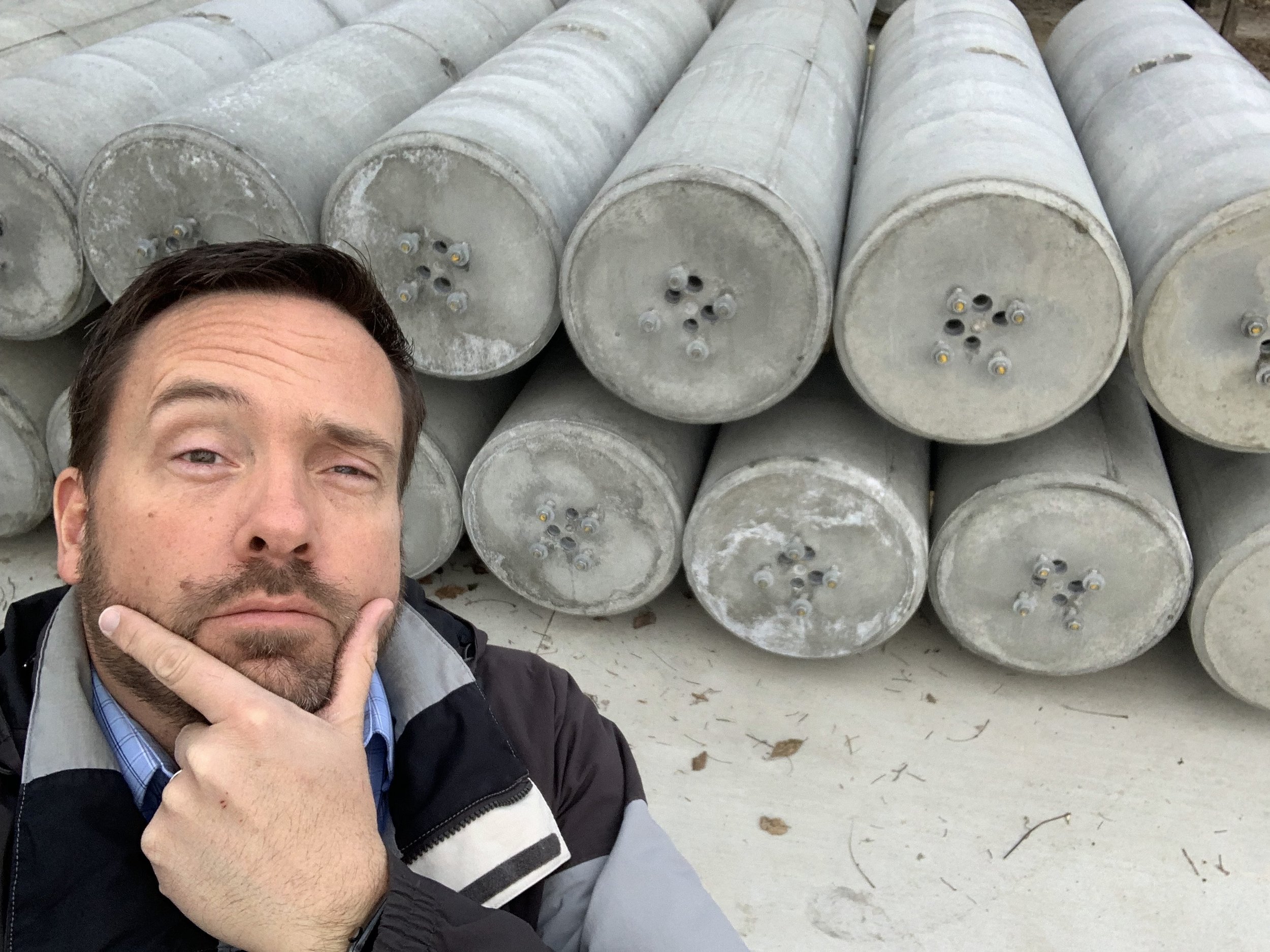 A man with a thoughtful expression, resting his chin on his hand, standing in front of a pile of large concrete or metal pipes arranged horizontally.