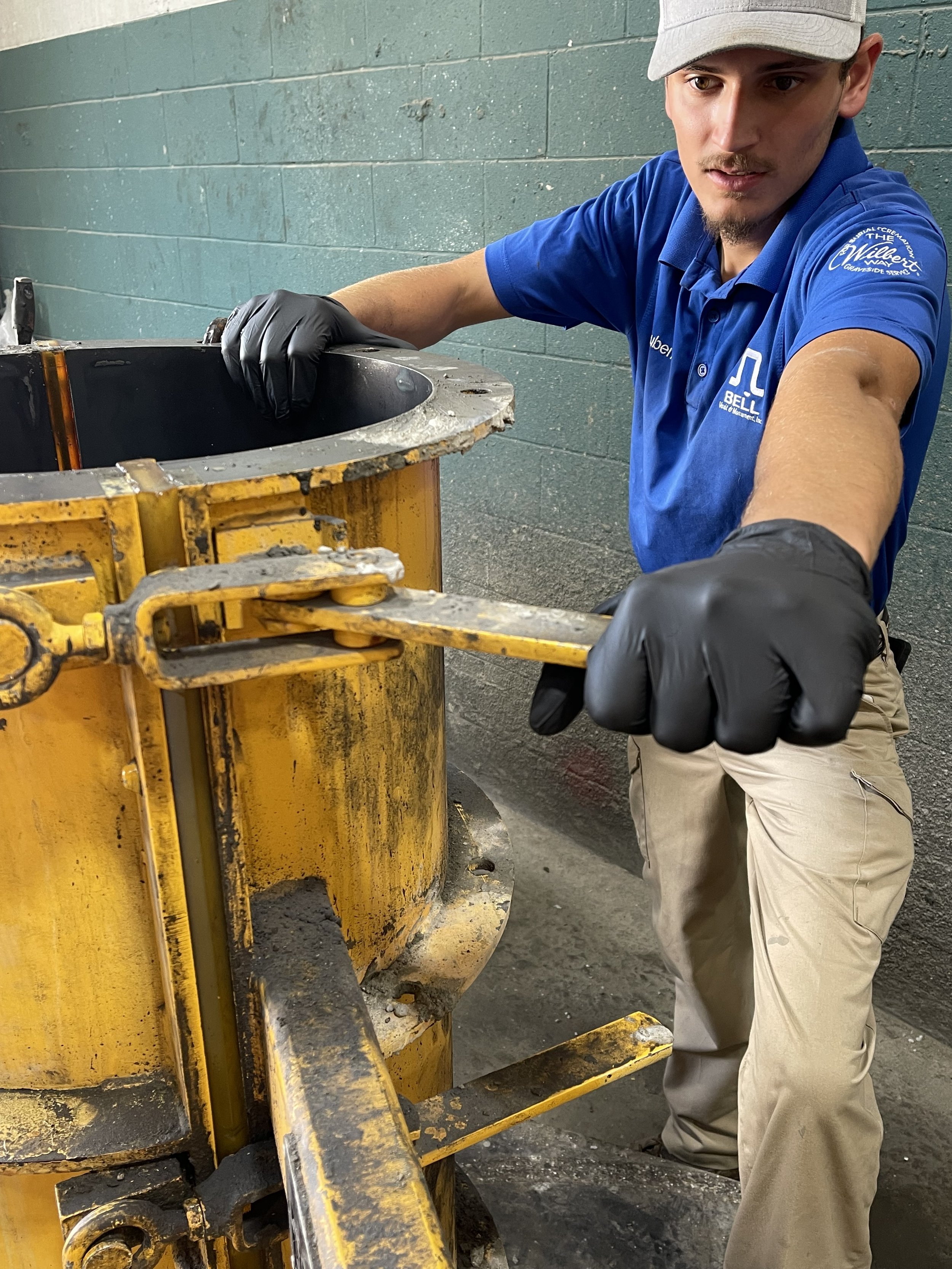 A man wearing a gray cap, blue polo shirt, and black gloves is working with industrial equipment in a workshop with teal brick walls.