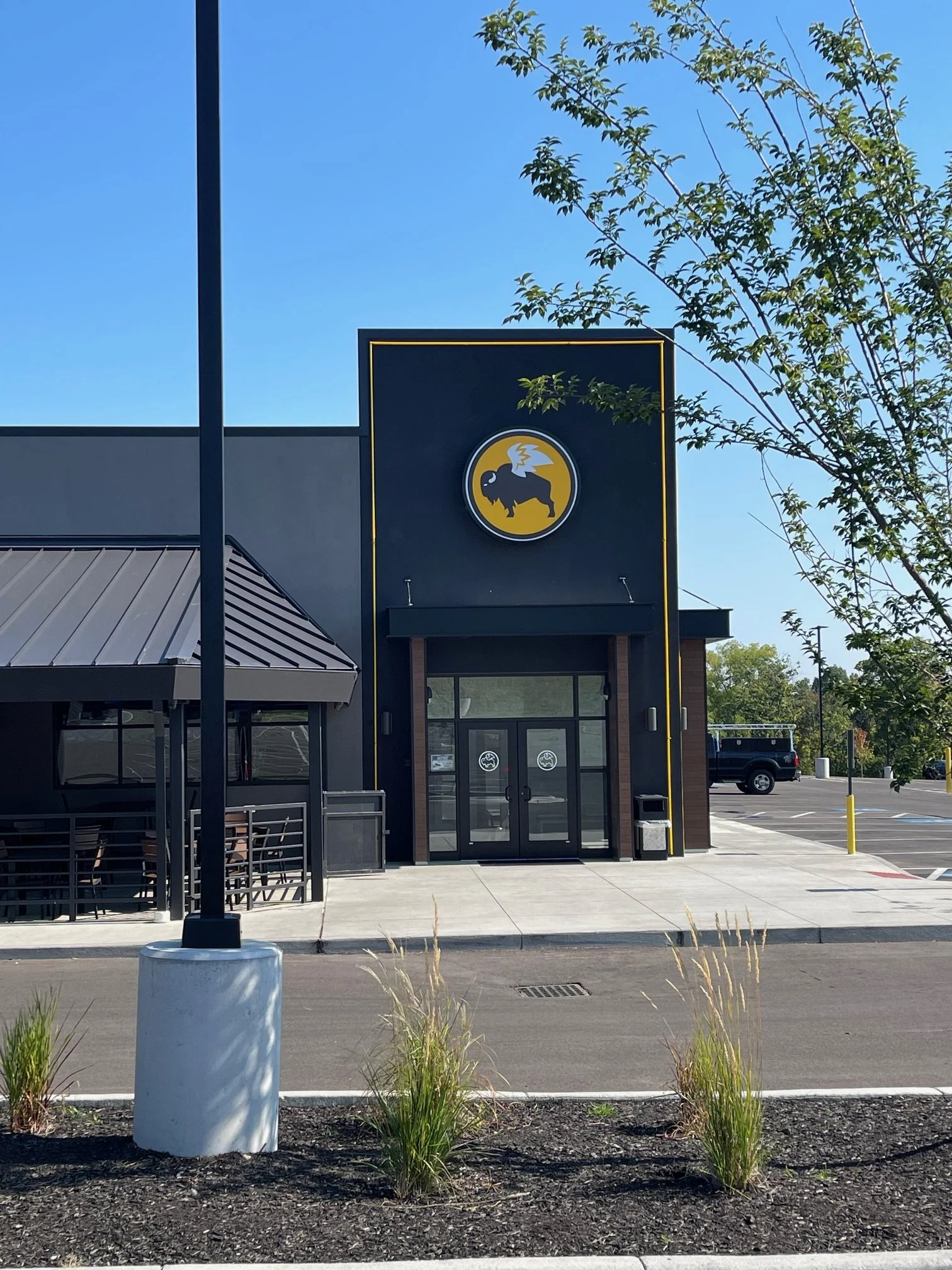 Exterior of a building with a black facade and large logo of a black buffalo with white wings inside a yellow circle, possibly for a restaurant or retail store, on a clear day with blue sky and some trees.