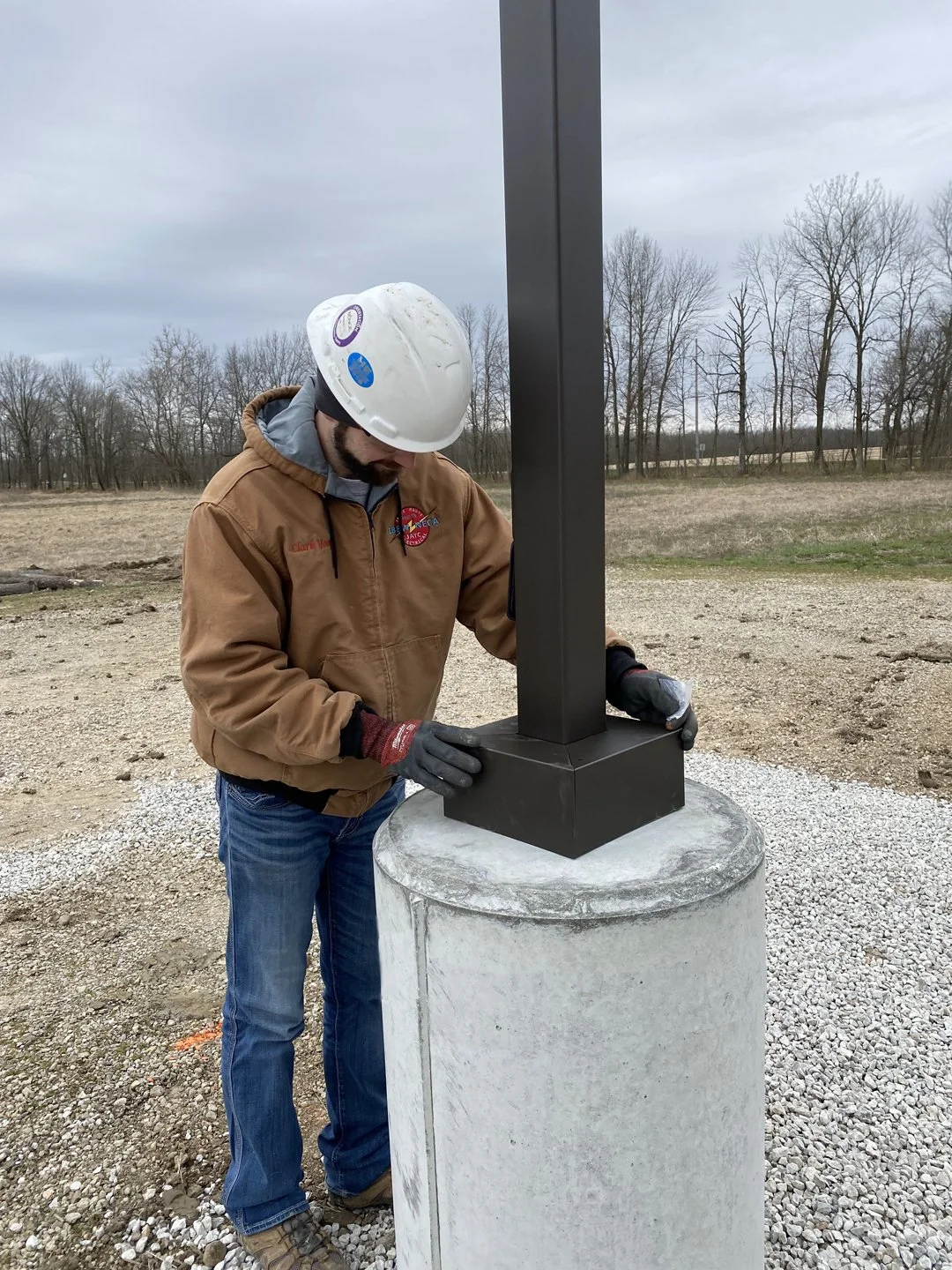 A worker wearing a white safety helmet, brown jacket, and gloves inspecting a black metal post mounted on a concrete pedestal outdoors in a rural area with bare trees in the background.