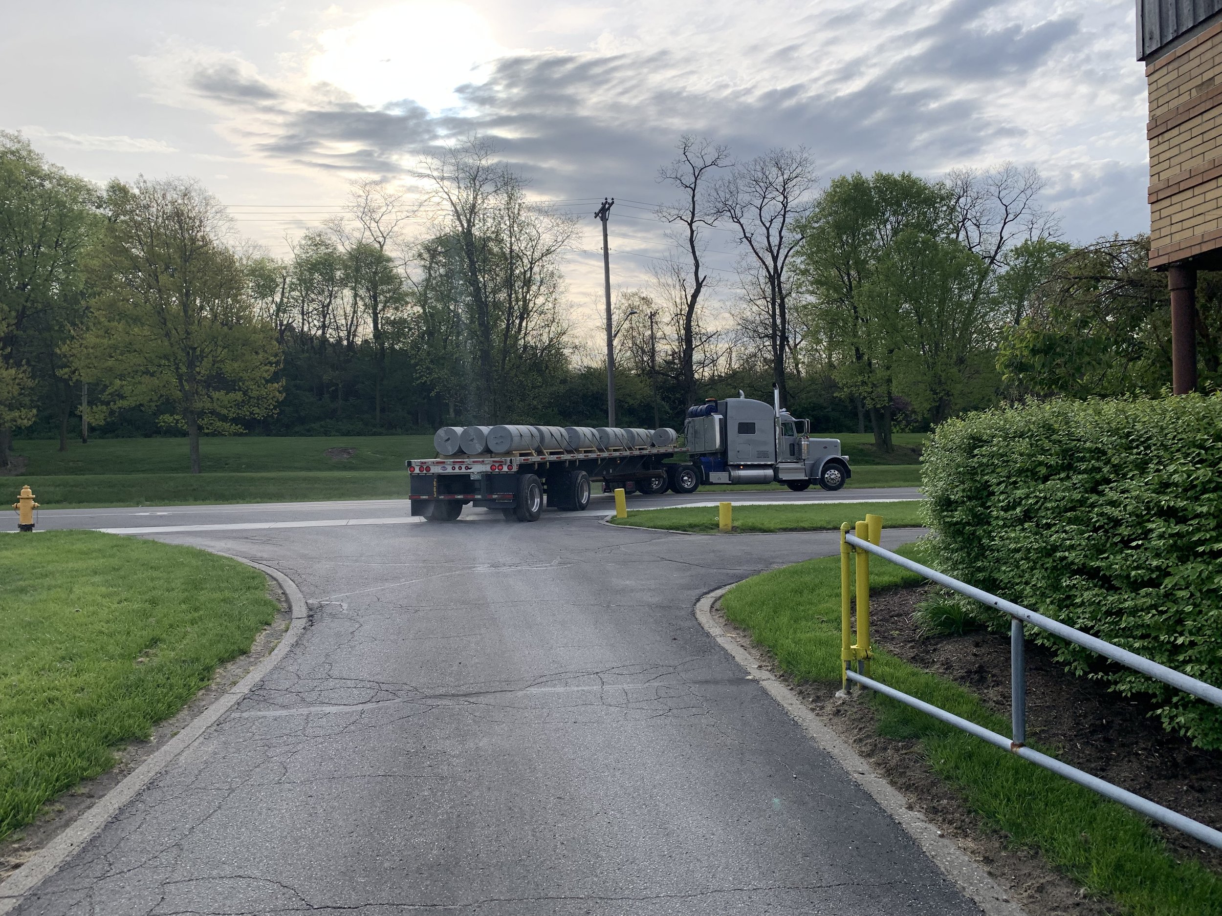 A semi-truck with a flatbed trailer carrying cylindrical water tanks is crossing a small curve in a driveway, with grass, trees, and a cloudy sky in the background.