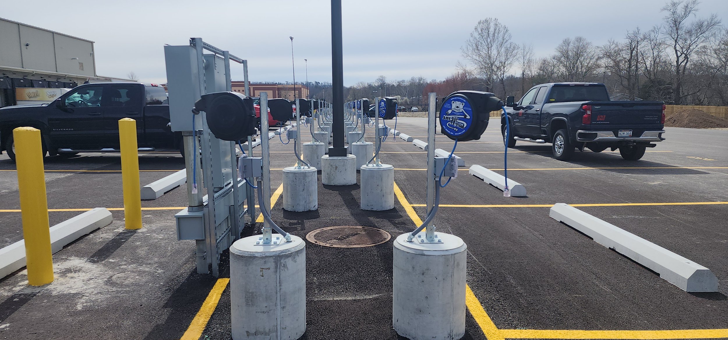 Empty parking lot with electric vehicle charging stations in foreground, two trucks parked in background, leafless trees and cloudy sky