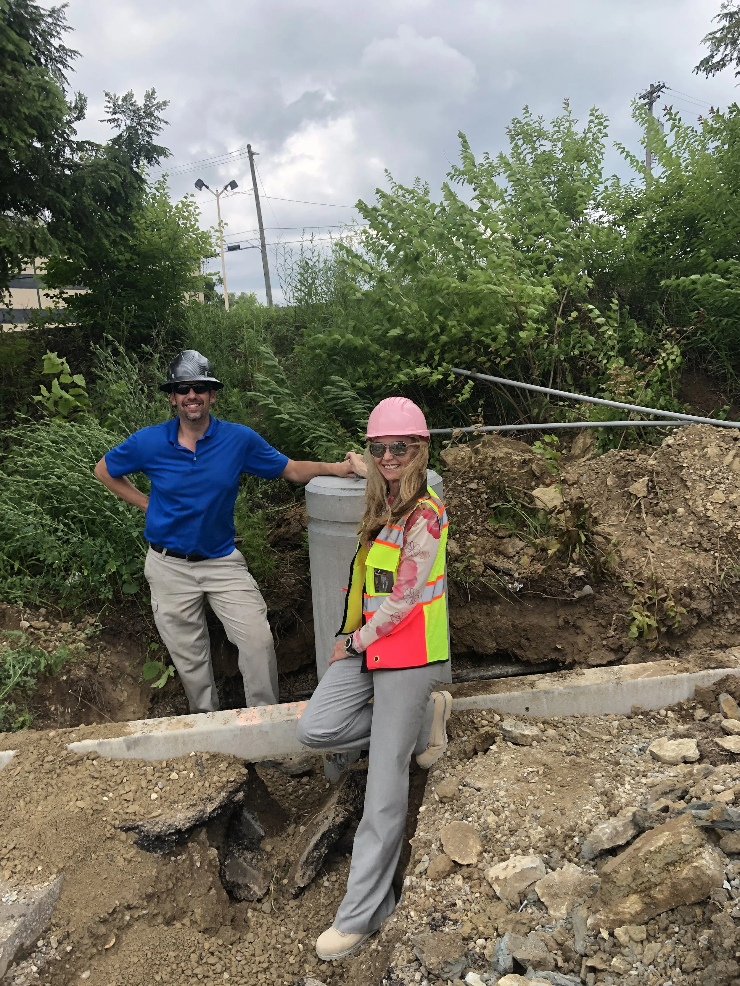 Two construction workers, a man in a black helmet and sunglasses and a woman in a pink helmet and sunglasses, are at a construction site with dirt and rocks. They are standing next to a large utility pole and smiling at the camera, with green foliage and a cloudy sky in the background.