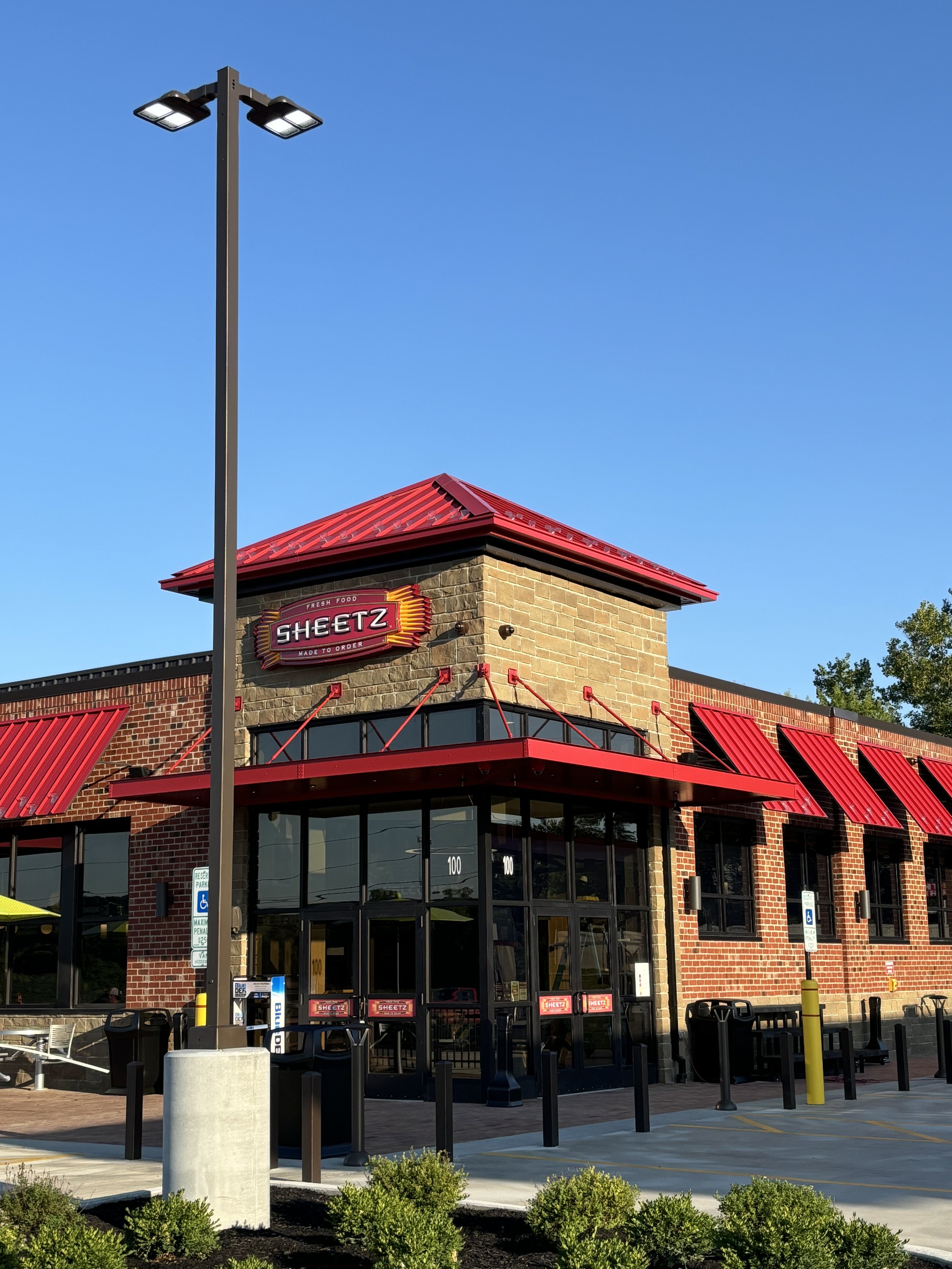 Exterior of a Sheetz convenience store with red awnings, brick and stone facade, and a parking lot with a light pole and small bushes in the foreground.